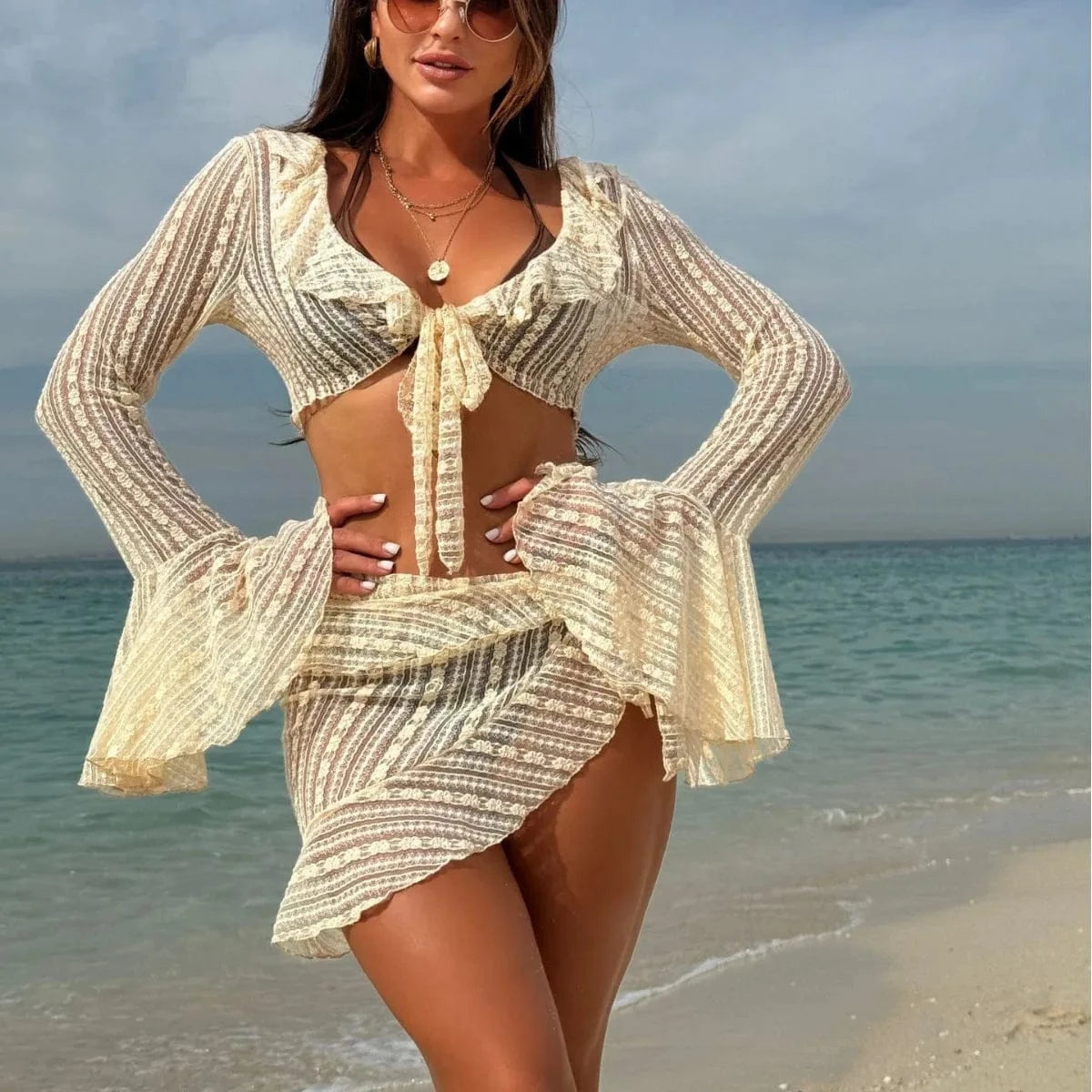 A woman stands on a sandy beach wearing a cream-colored, two-piece lace set consisting of a long-sleeved, ruffled crop top with a front tie and a matching asymmetrical wrap-style mini skirt.