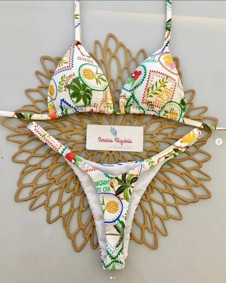 A white triangle bikini set featuring a colorful postage stamp-style tropical print with illustrations of fruits and plants, displayed on a decorative golden laser-cut placemat.