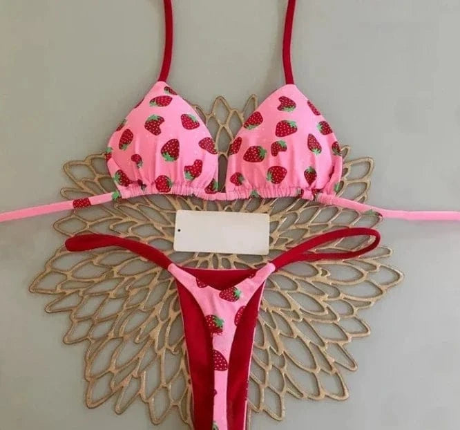 A pink two-piece bikini featuring a small strawberry pattern, with red straps and trim, laid flat on a decorative gold-toned circular placemat.