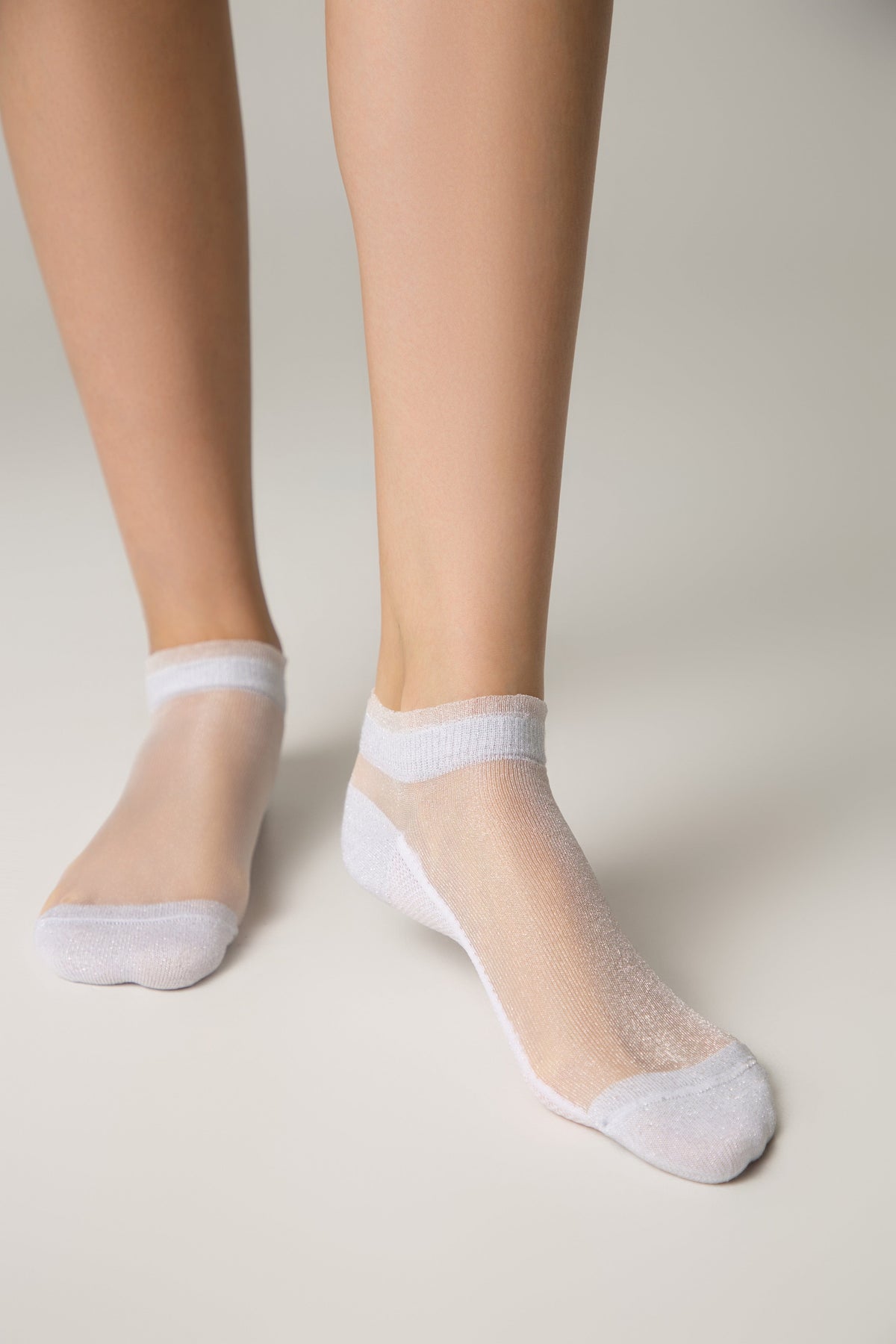A pair of sheer white ankle socks with a subtle shimmer worn on a person's feet against a light grey background.