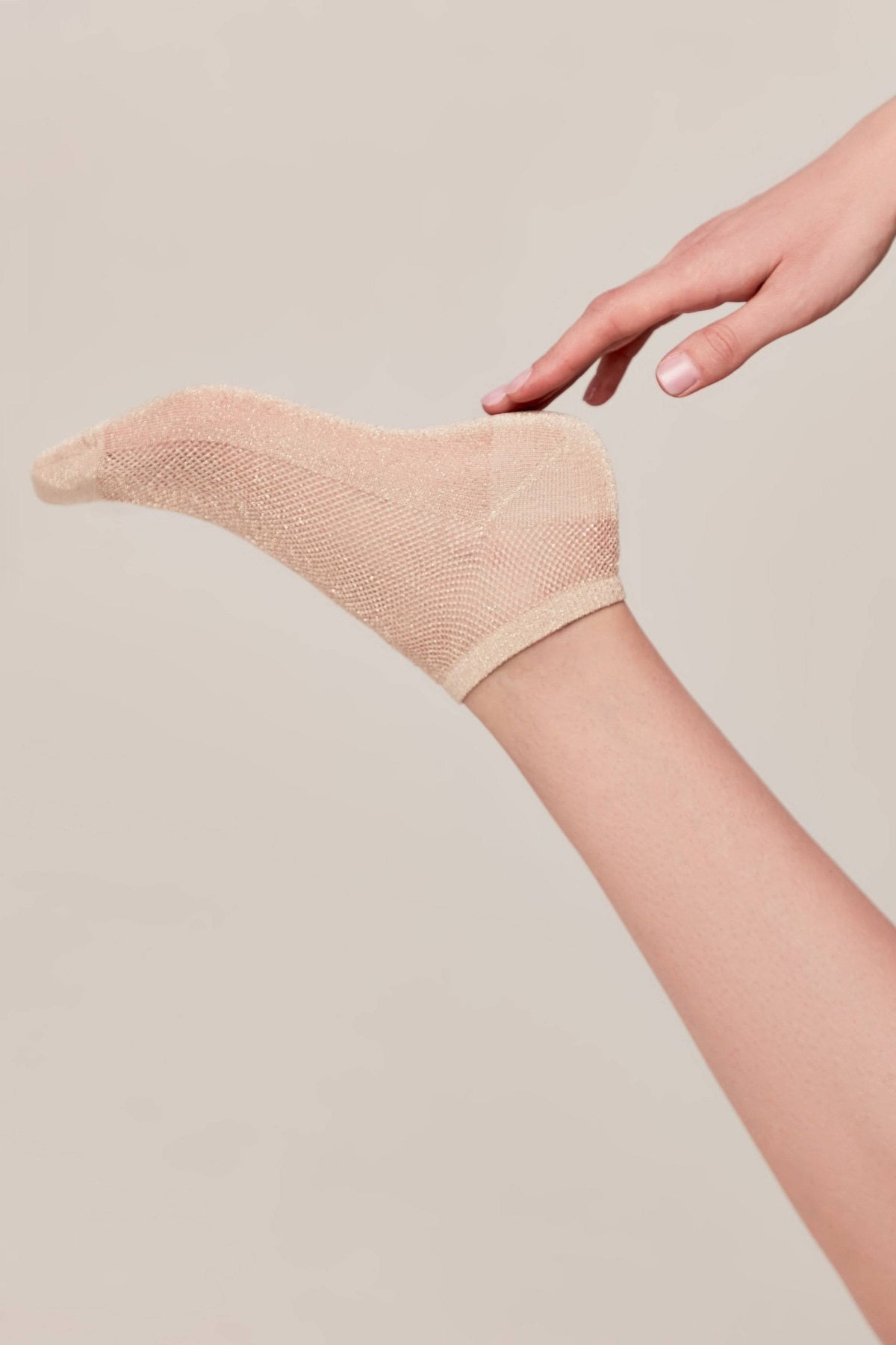 A beige glittery ankle sock worn on a foot, with a hand gently touching the heel against a plain neutral background.