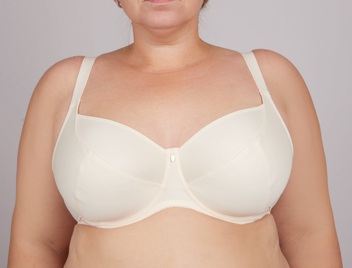 A beige full-coverage underwire bra worn by a person, featuring smooth cups, wide adjustable straps, and a small central charm detail.