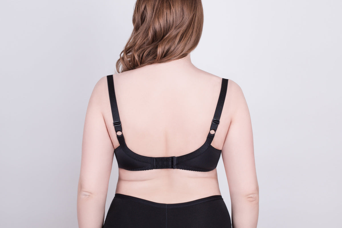 A black bra viewed from the back on a person with shoulder-length wavy brown hair against a plain light gray background.