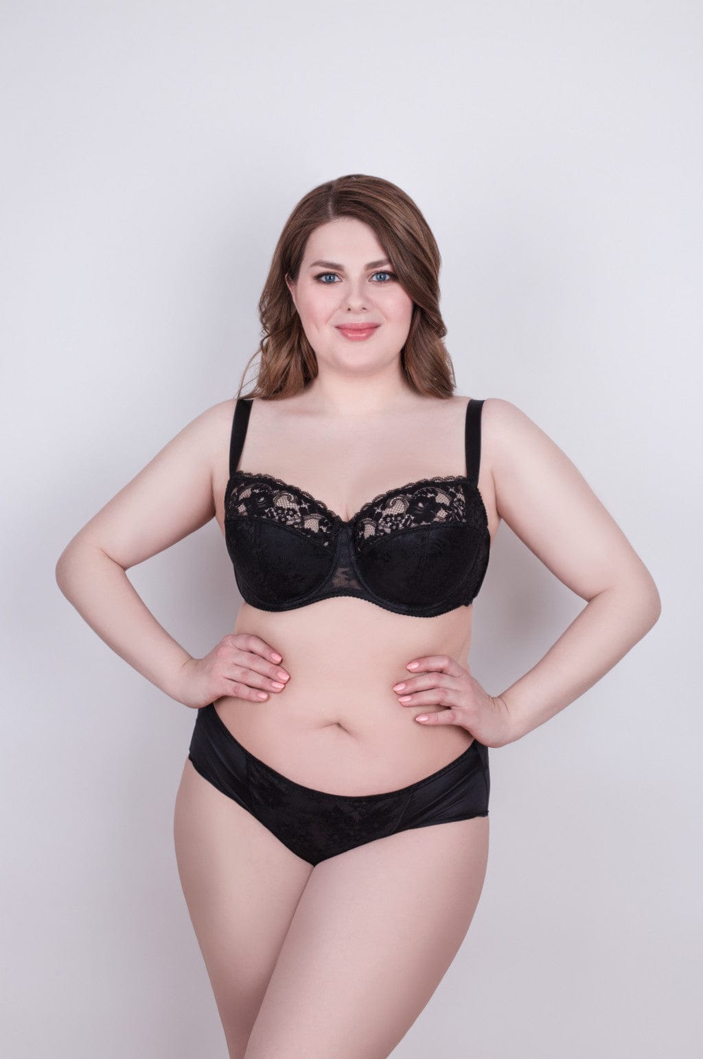 A woman models a black two-piece lingerie set with lace detailing on the bra cups and a solid black panty against a plain light gray background.