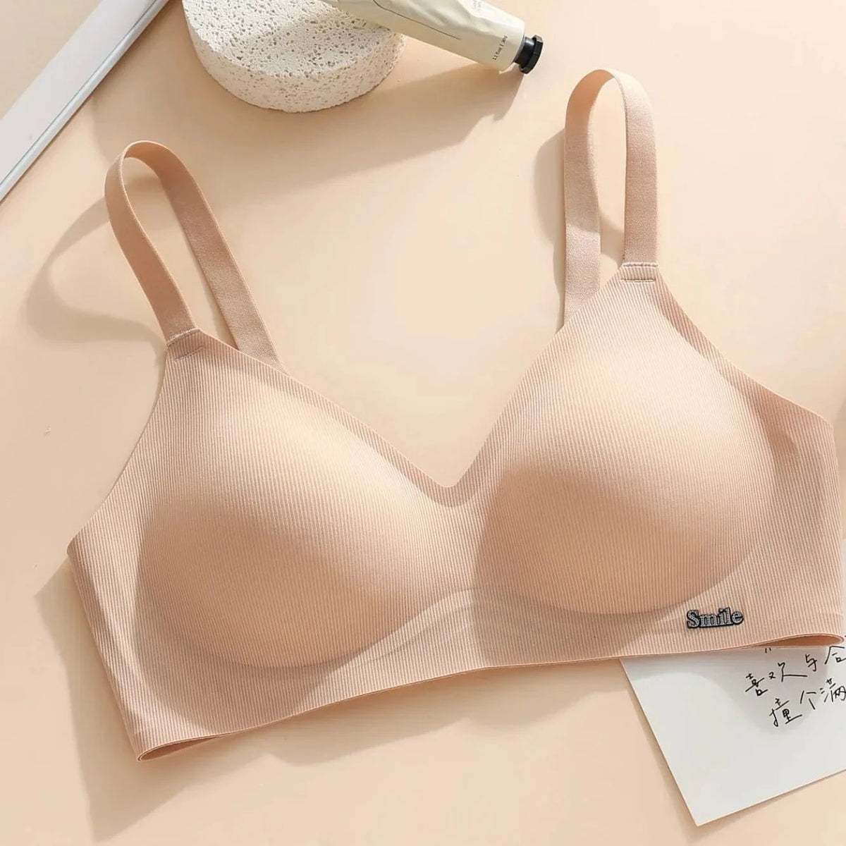 A beige ribbed-texture wireless bra with adjustable straps sits on a light surface next to a tube of cream and a card with handwritten text.