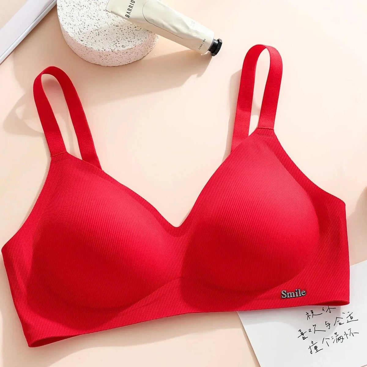 A vibrant red wireless bra with ribbed fabric features wide straps, a deep neckline, and a small silver "Smile" charm on the lower right side, placed on a light surface next to a white tube of cream and a circular textured prop.