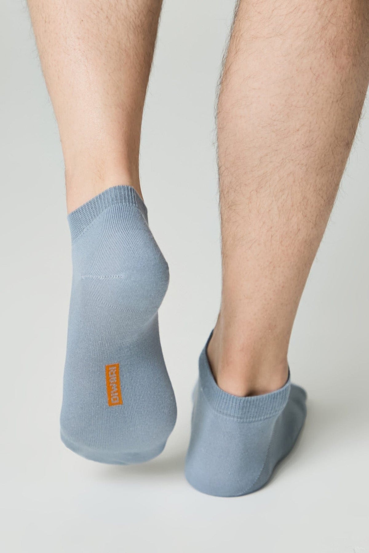 A pair of light blue ankle socks are worn on human feet, featuring a small orange vertical logo on the heel of the left sock.