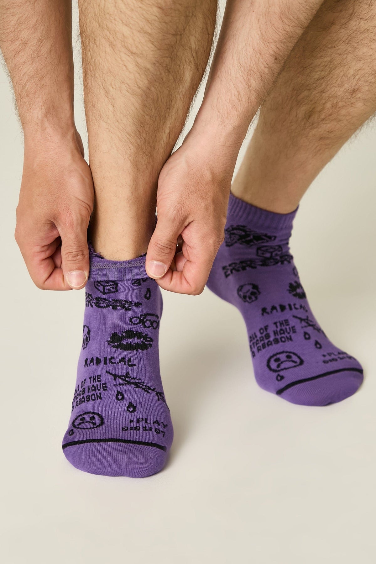 A person pulls on a pair of purple ankle socks featuring a black printed design with various hand-drawn style graphics, including a sad face, a skull, raindrops, and text reading RADICAL and all of the stars have a reason.