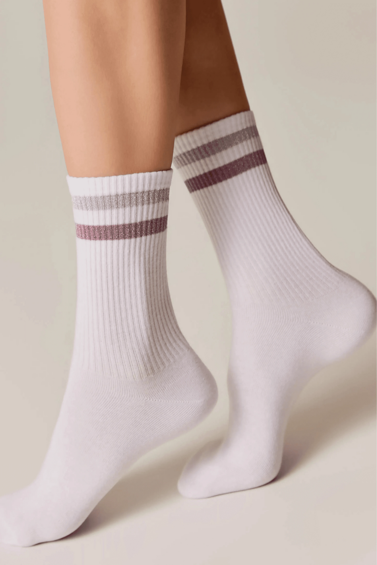 A pair of white crew socks featuring ribbed texture and horizontal pink and silver metallic stripes at the cuffs, worn on legs against a plain background.