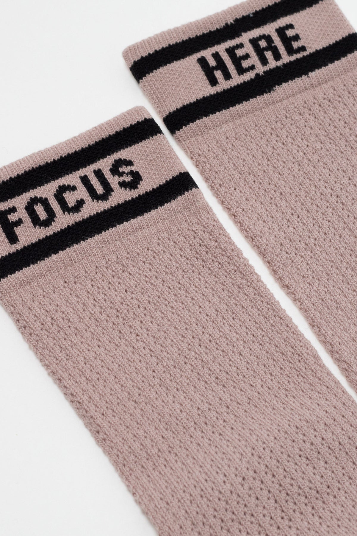 A pair of dusty pink, textured crew socks with a black striped cuff featuring the word FOCUS on one sock and HERE on the other in bold black lettering.