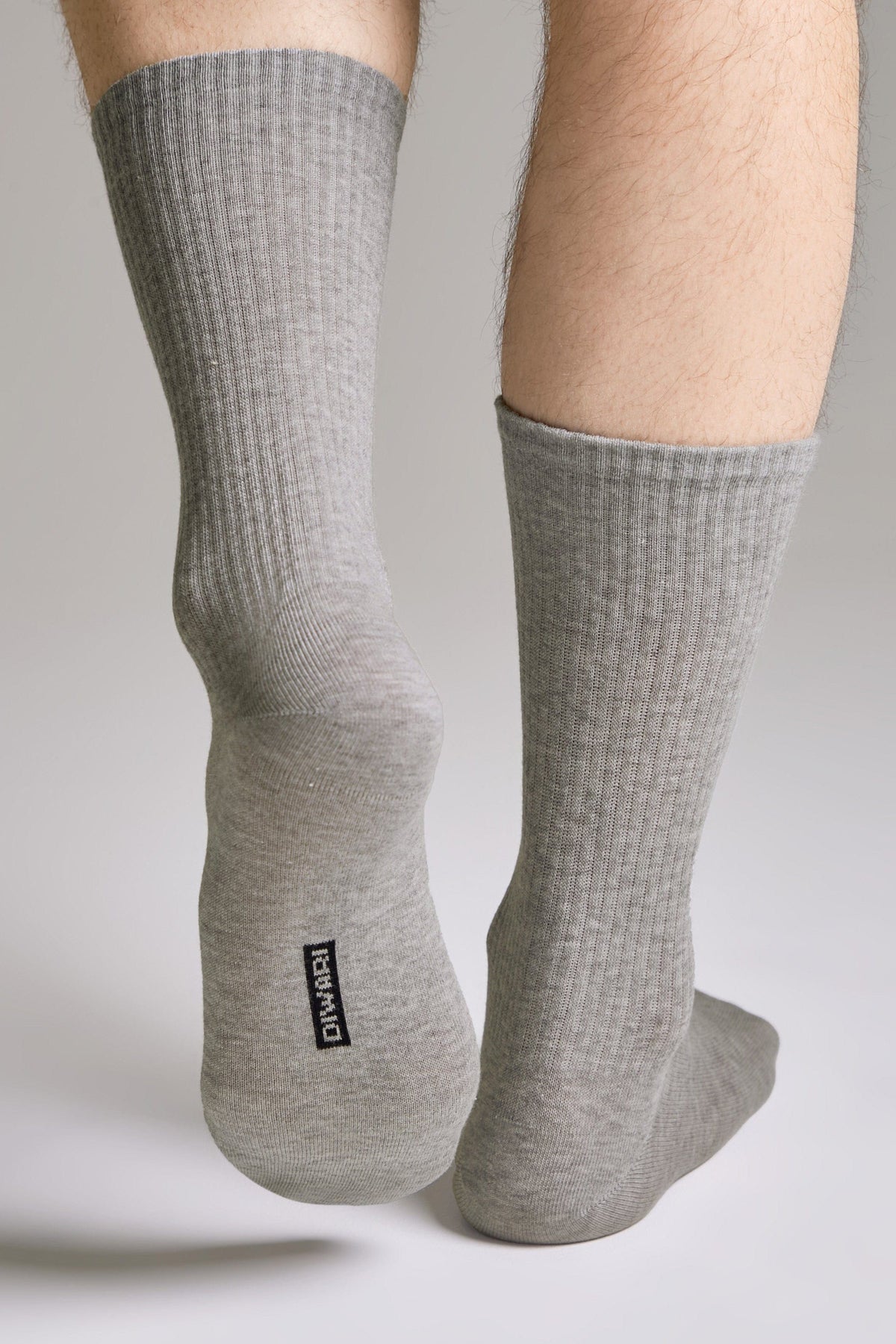 A pair of grey ribbed crew socks worn on a person's feet, featuring a small rectangular black label with white lettering on the sole.