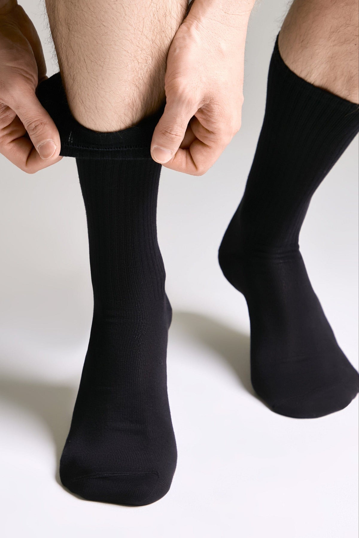 A pair of solid black crew socks on a person's feet, with hands shown pulling the cuff of the left sock upward against a plain light gray background.