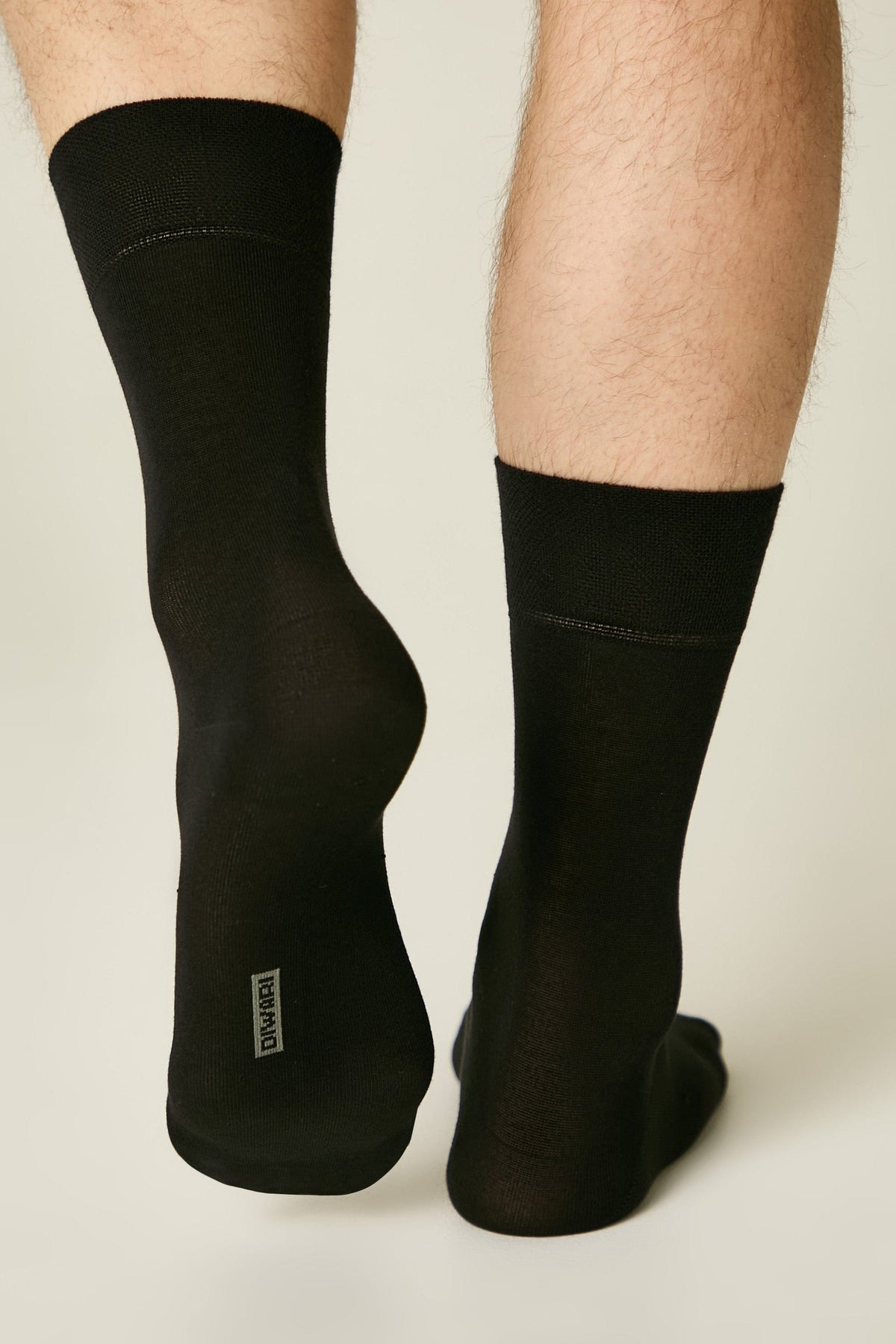 A pair of black crew socks worn on a person's lower legs, showing a small grey rectangular logo on the heel of one sock against a light grey background.