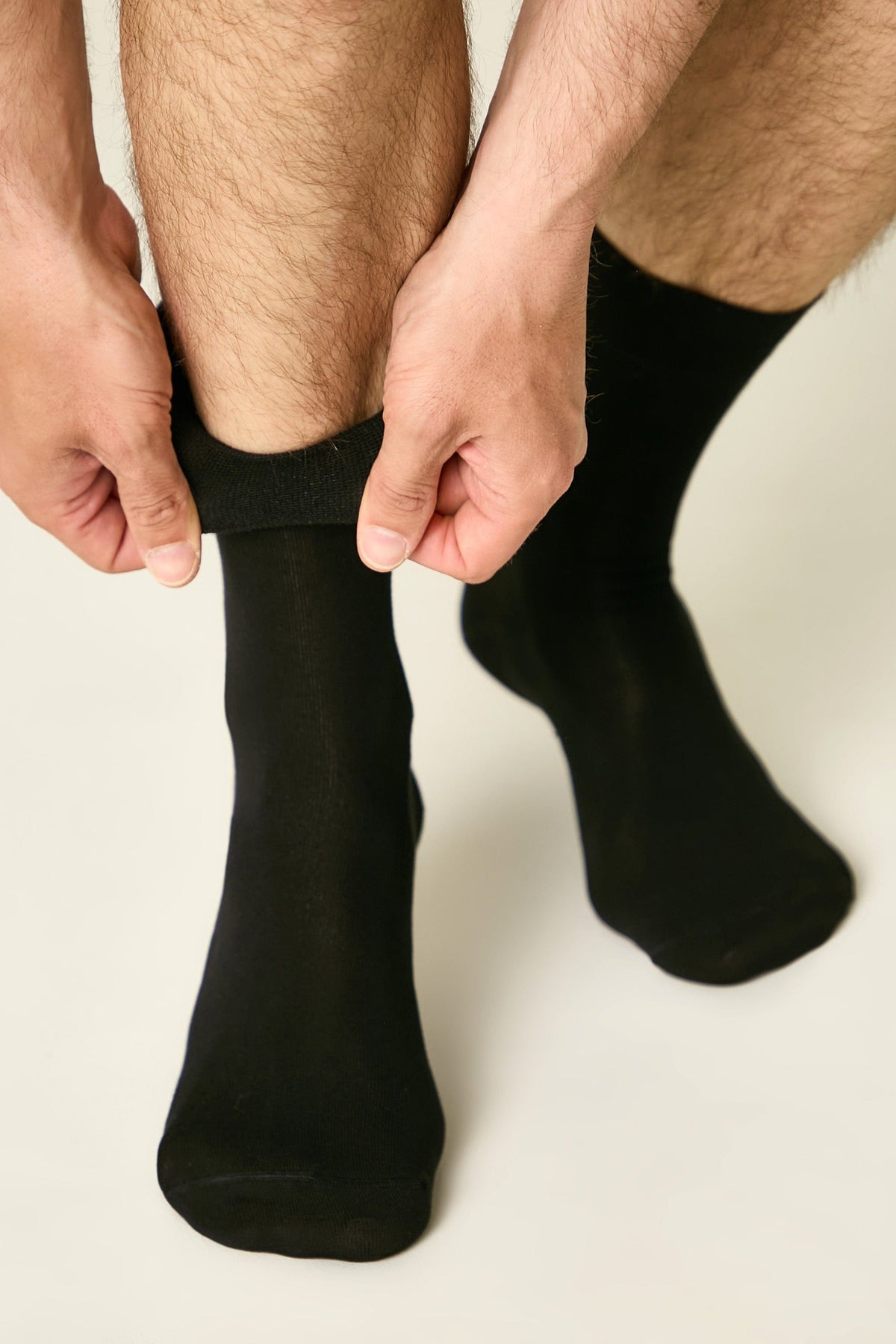 A pair of plain black crew socks worn on a person's feet, with hands shown pulling the cuff of one sock up.