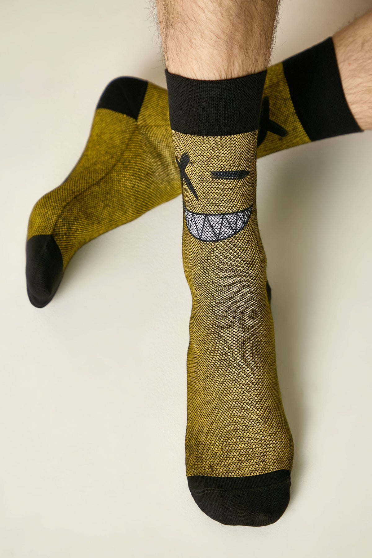 A pair of yellow crew socks featuring a black, winking cartoon face design with a jagged grin on a textured fabric background, worn on a person's lower legs against a neutral backdrop.