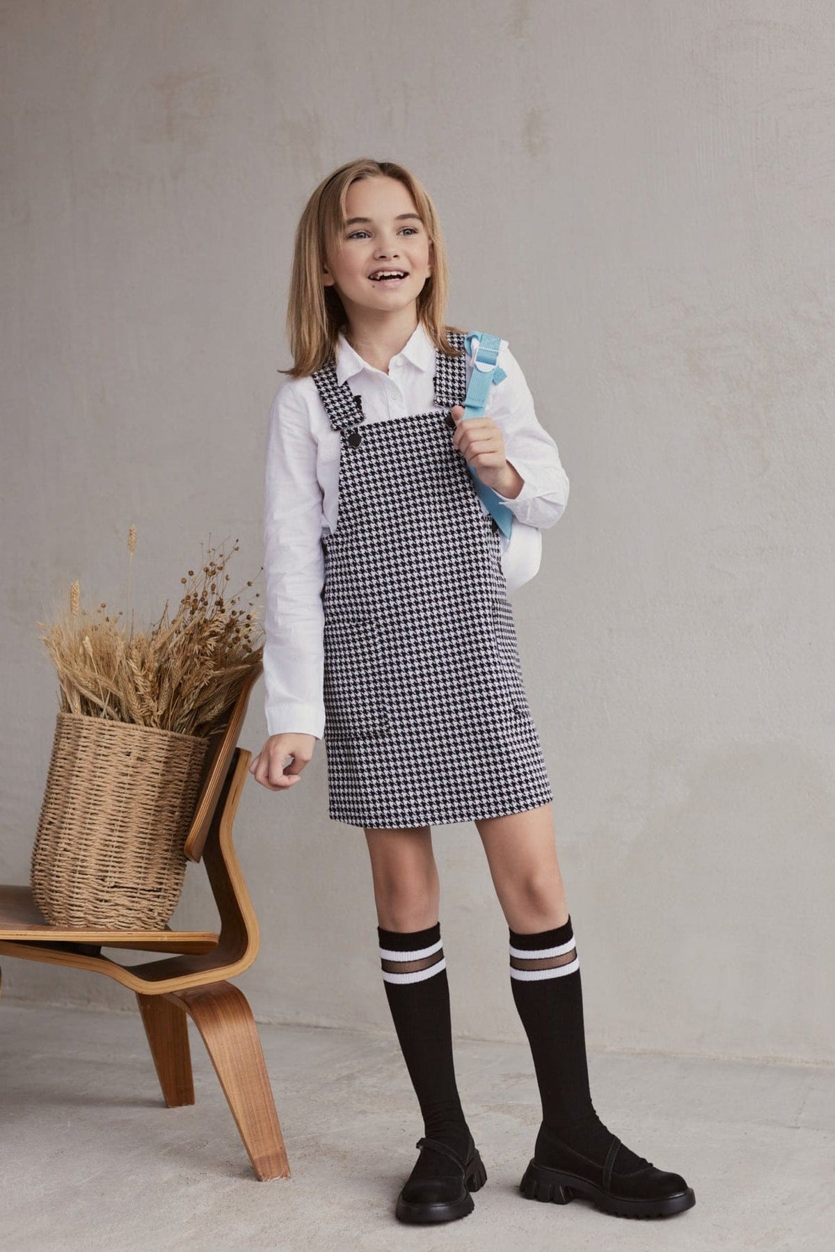 A young girl stands wearing a white long-sleeved collared shirt under a black and white houndstooth pinafore dress, paired with black knee-high socks featuring white stripes and black platform shoes, standing next to a chair holding a woven