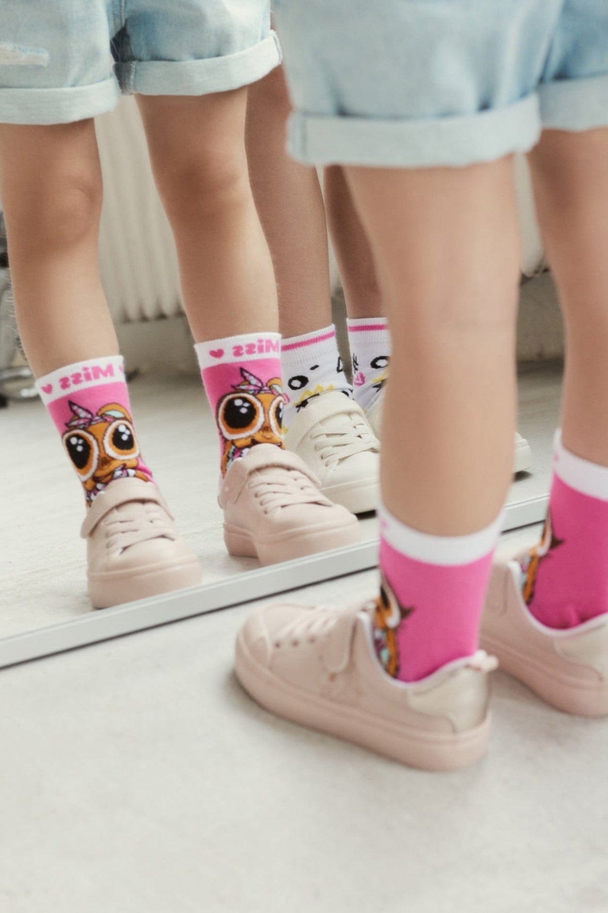 Pink socks featuring a cartoon owl character are worn by a child in light blue denim shorts and light pink sneakers, reflected in a mirror on the floor.