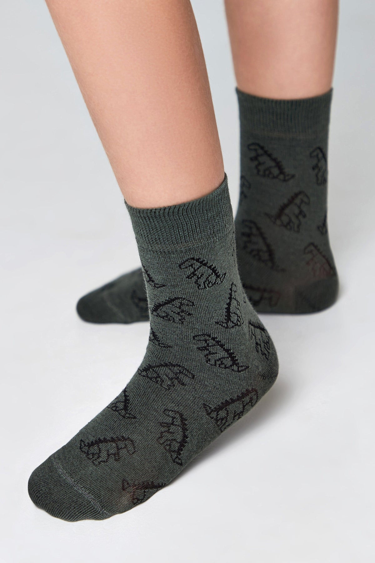 Olive green crew-length socks featuring a repeating dark brown dinosaur pattern worn on a person's lower legs against a plain background.