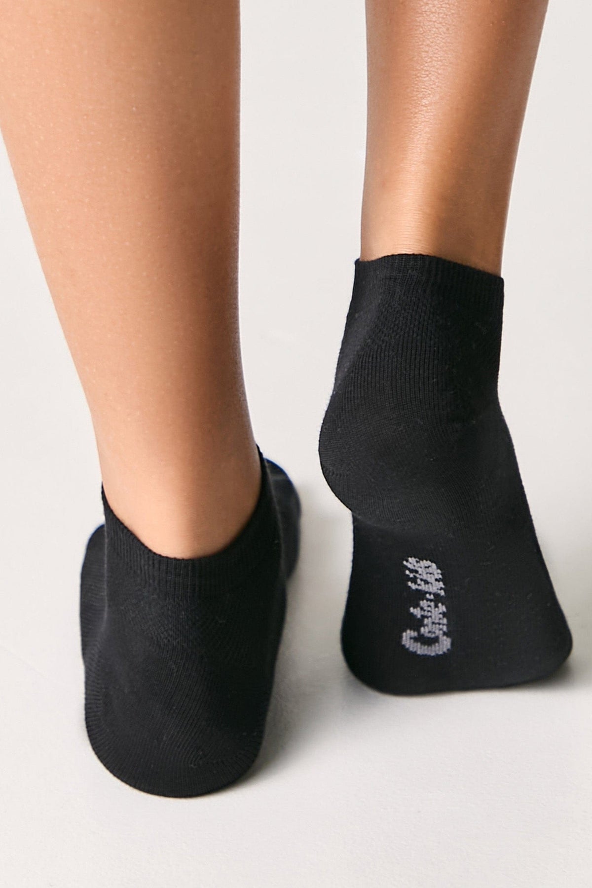A pair of black ankle socks worn on the feet, featuring a small white logo printed on the sole of the right sock.