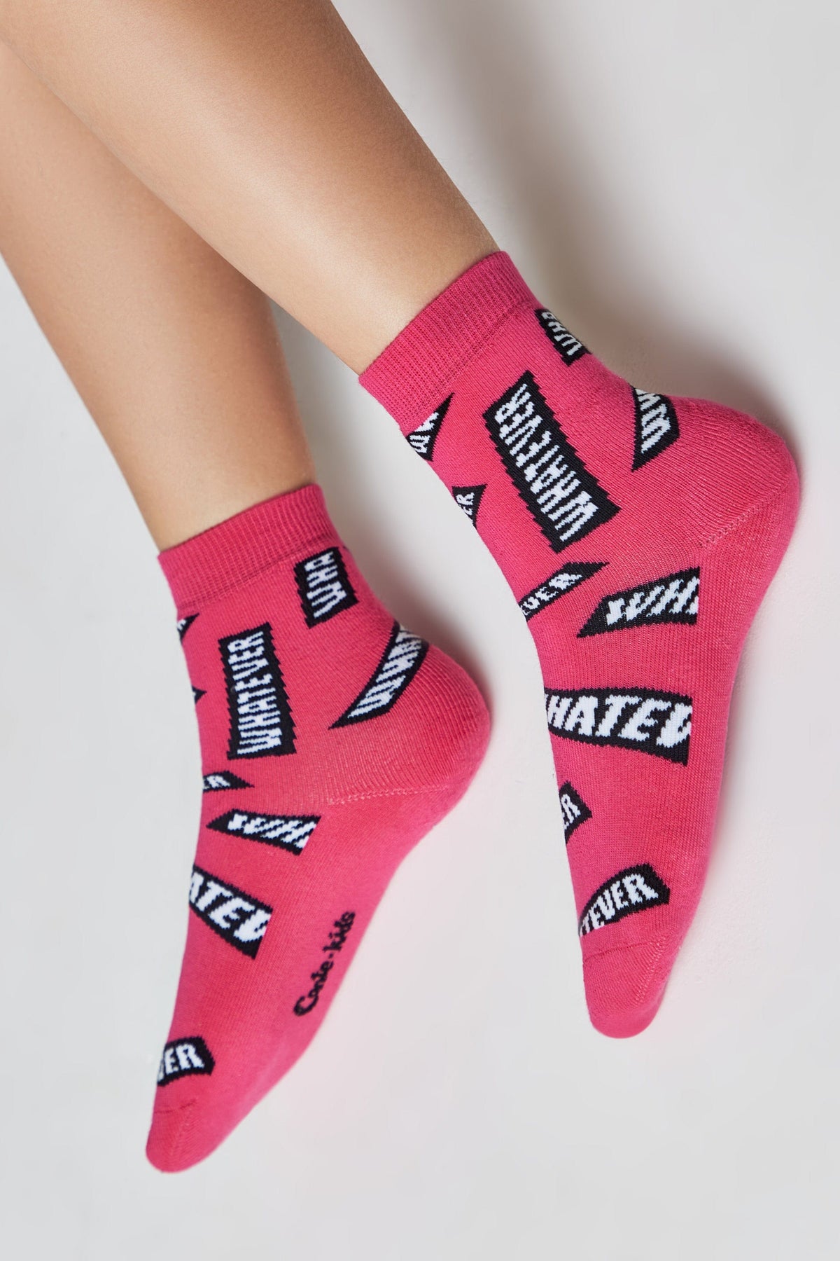 A pair of bright pink ankle socks featuring a repeating pattern of white text in black rectangles that reads "WHATEVER" worn on a person's feet against a plain white background.
