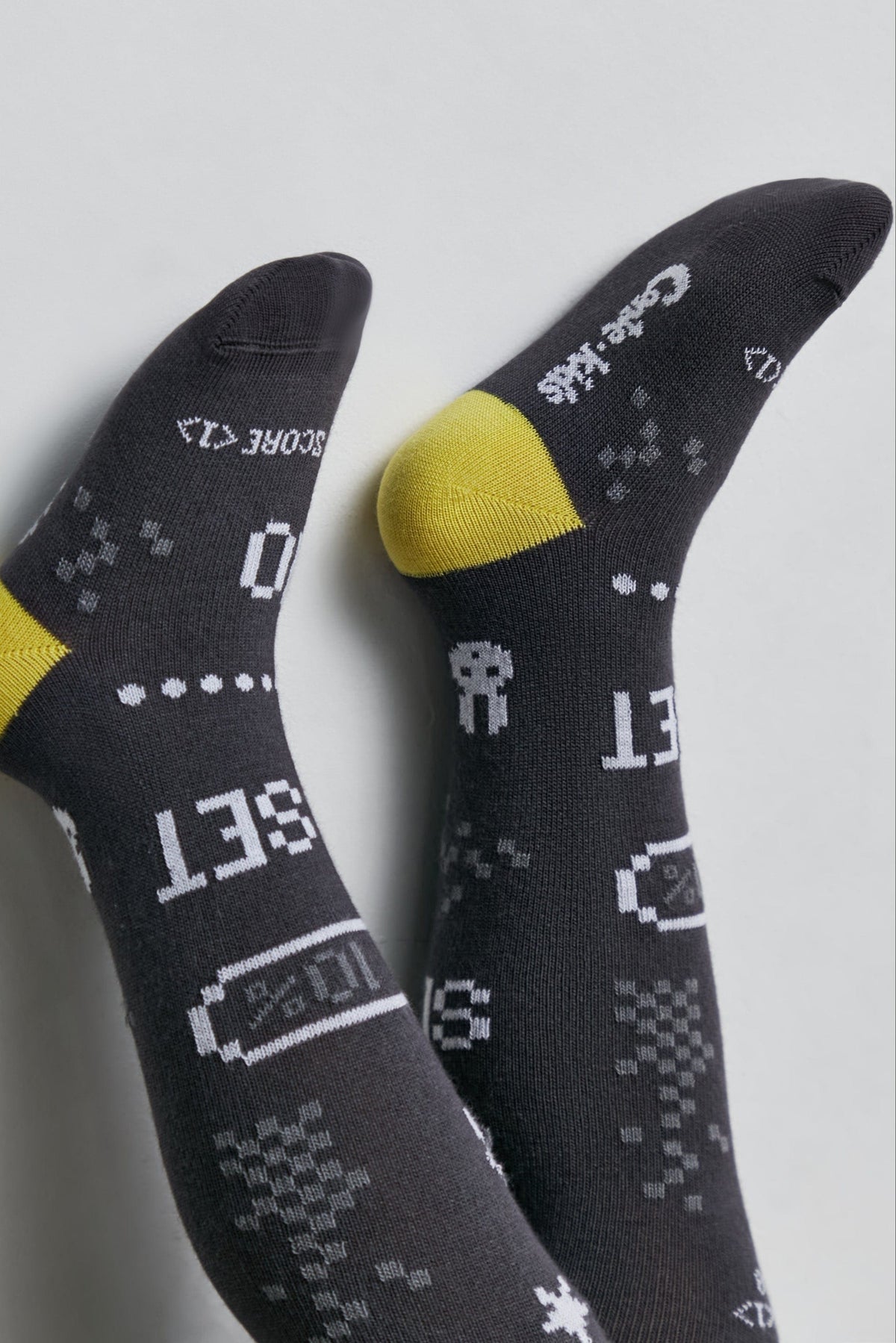 A pair of grey socks featuring white pixelated game-themed graphics like battery icons, text that reads "SET" and "SCORE", and yellow accents on the heels.