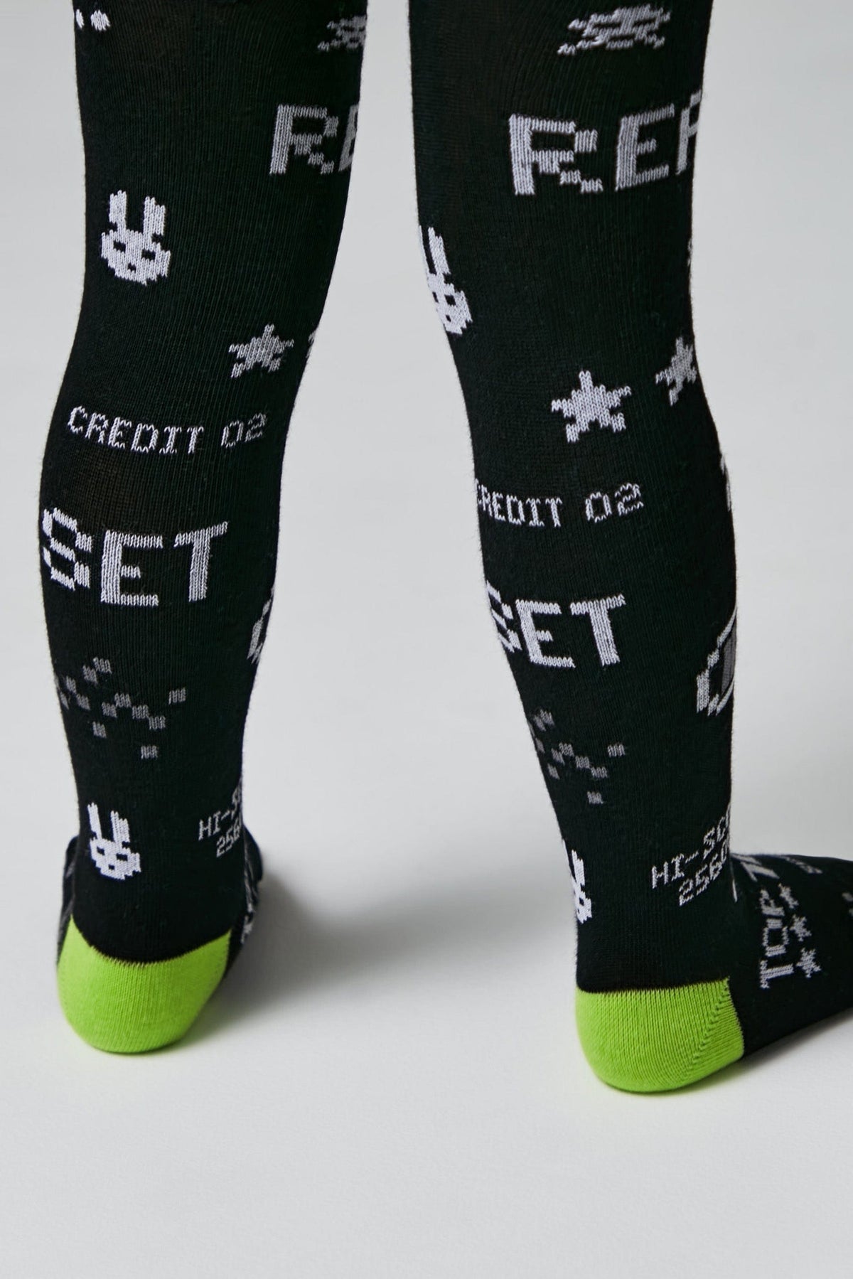 Black leggings featuring a repeating white pixelated pattern of bunnies, stars, and the words RESET and CREDIT 02, finished with bright green heels and toes.