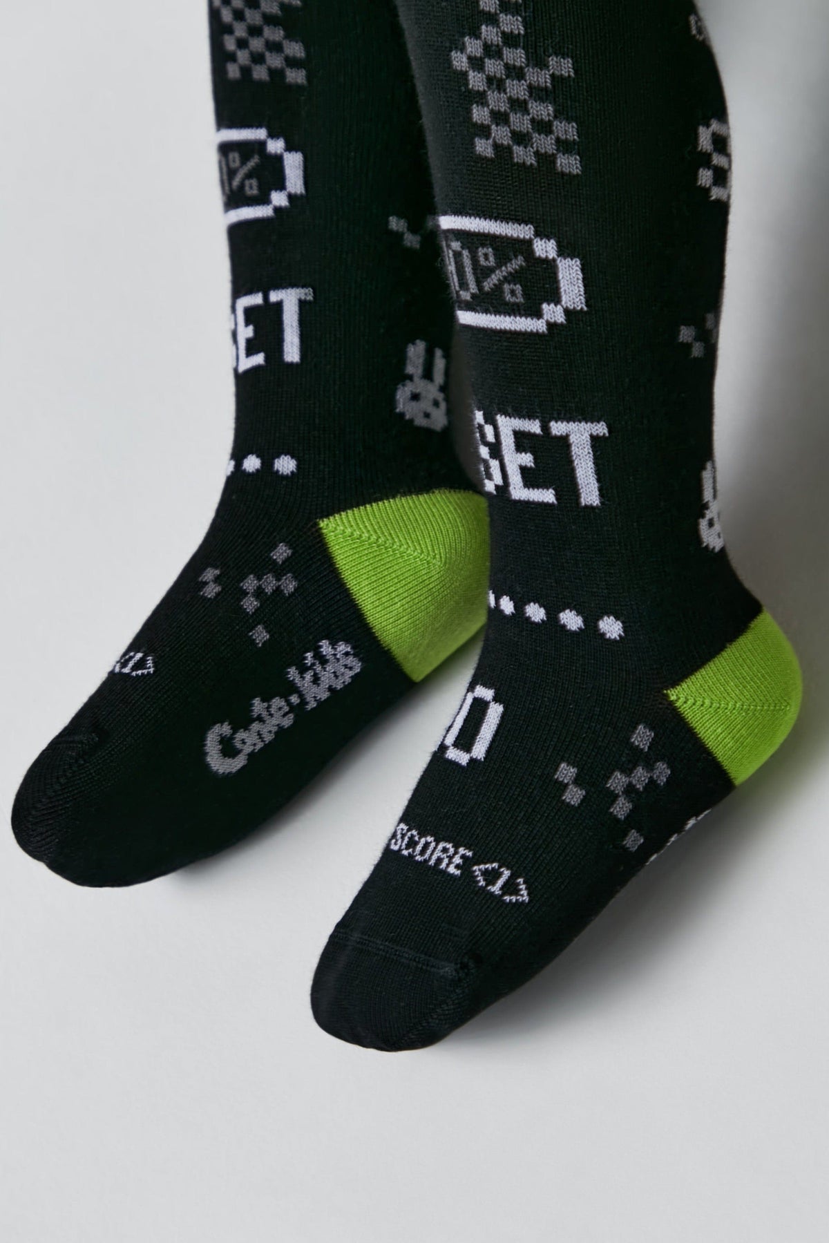 A pair of black children's tights featuring a pixelated design with the words SET and SCORE, a battery icon, and lime green accents on the heels and toes.