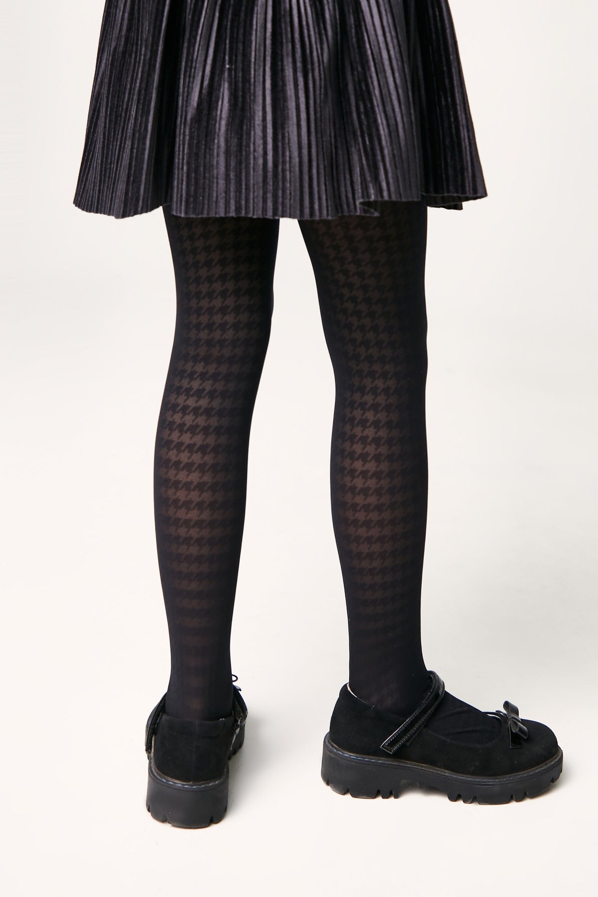 A pair of black opaque tights with a subtle houndstooth pattern worn with a black pleated skirt and black Mary Jane shoes with thick rubber soles.