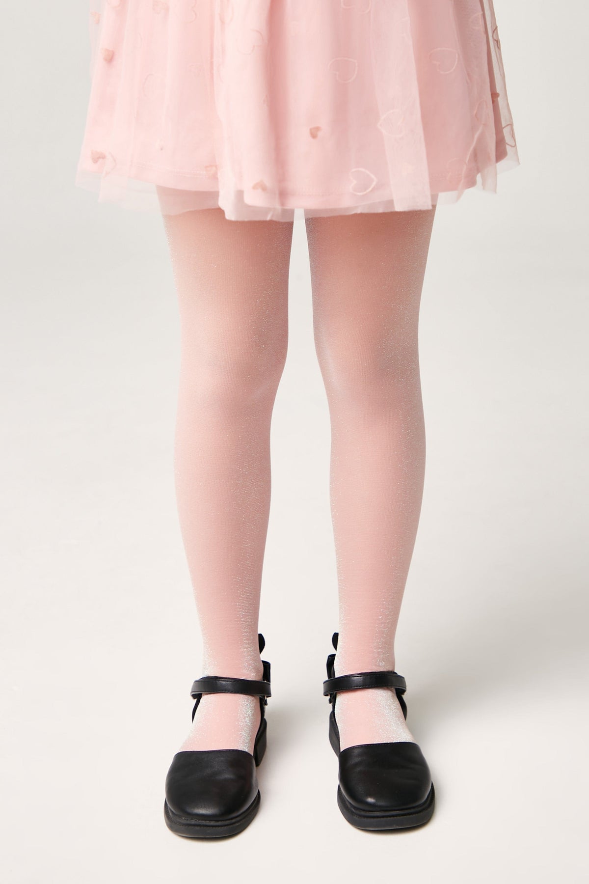 A child wears shimmering light pink tights paired with black Mary Jane shoes and a pale pink heart-patterned tulle skirt.