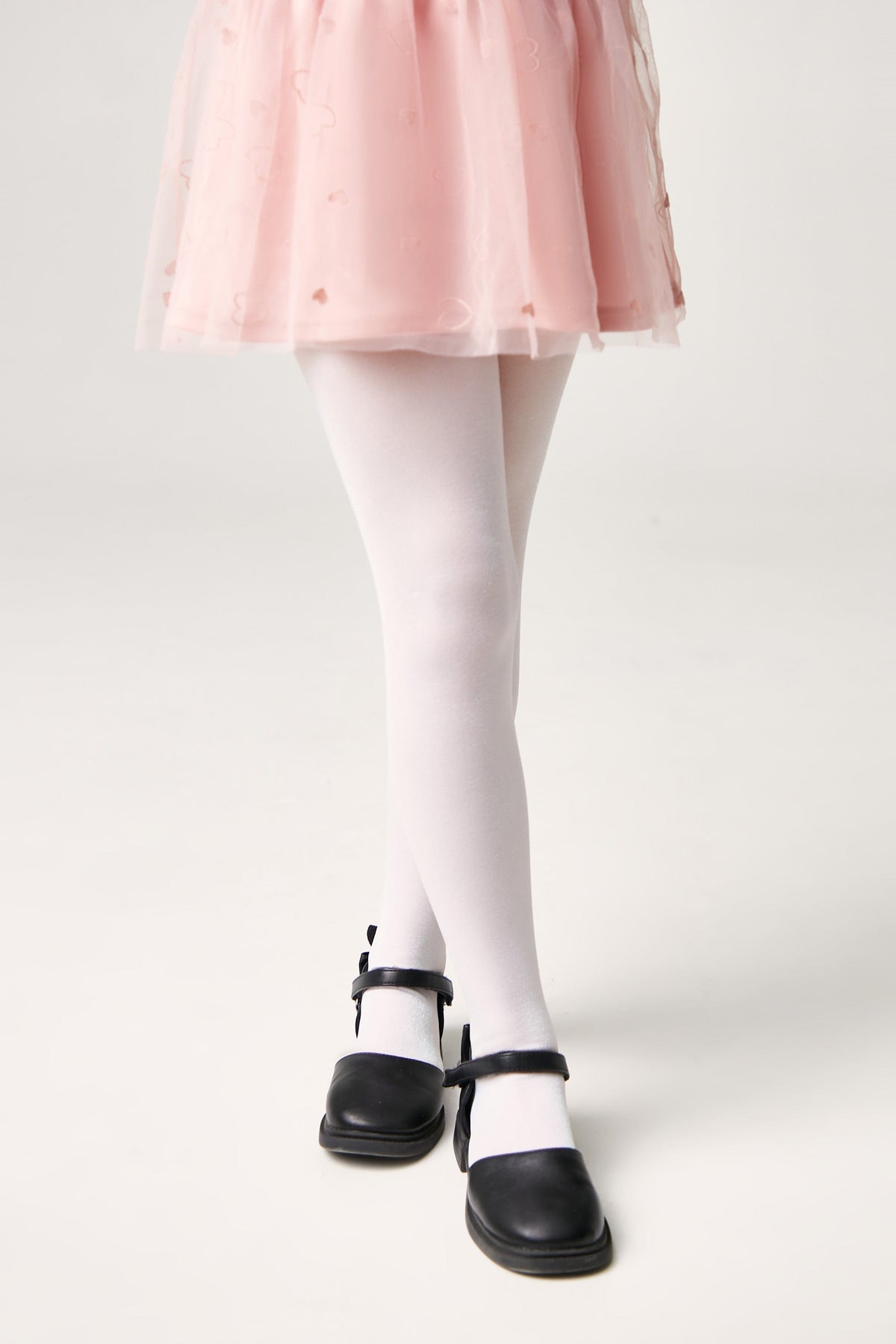 A pair of opaque white tights worn with a pink tulle skirt and black Mary Jane shoes against a white background.