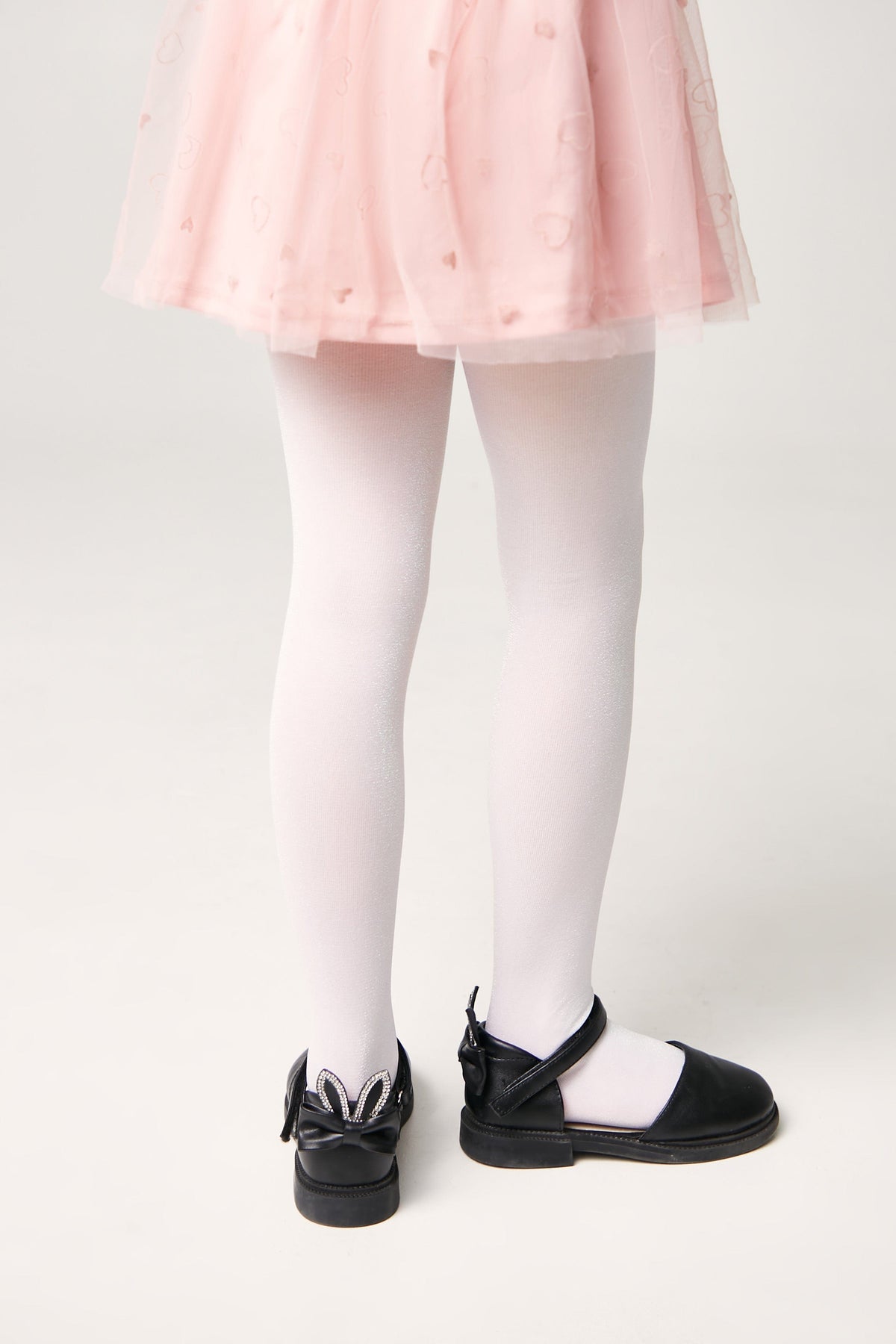 A pair of child's white tights paired with black Mary Jane shoes featuring a decorative bow and rabbit ears on the heel.