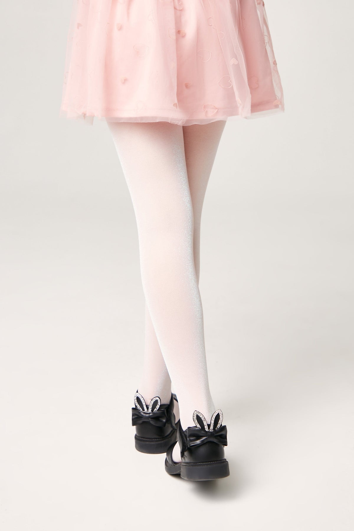 A child wears white sparkly tights and black ballet flats decorated with black bows and white rabbit ears with rhinestone detailing, paired with a pale pink tulle skirt.