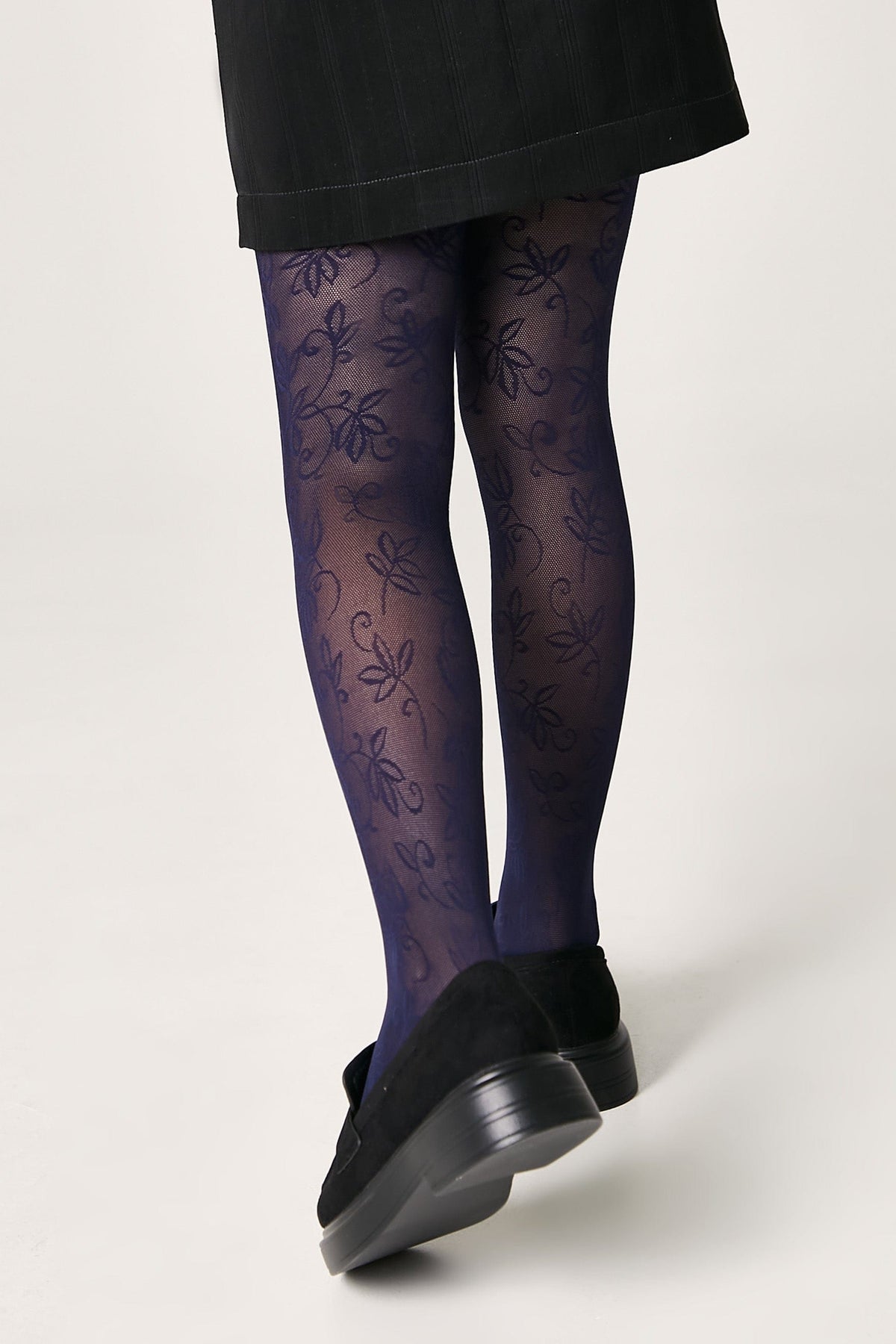 A person wears sheer, navy blue patterned tights with a delicate floral design paired with black loafers and a dark skirt against a plain background.