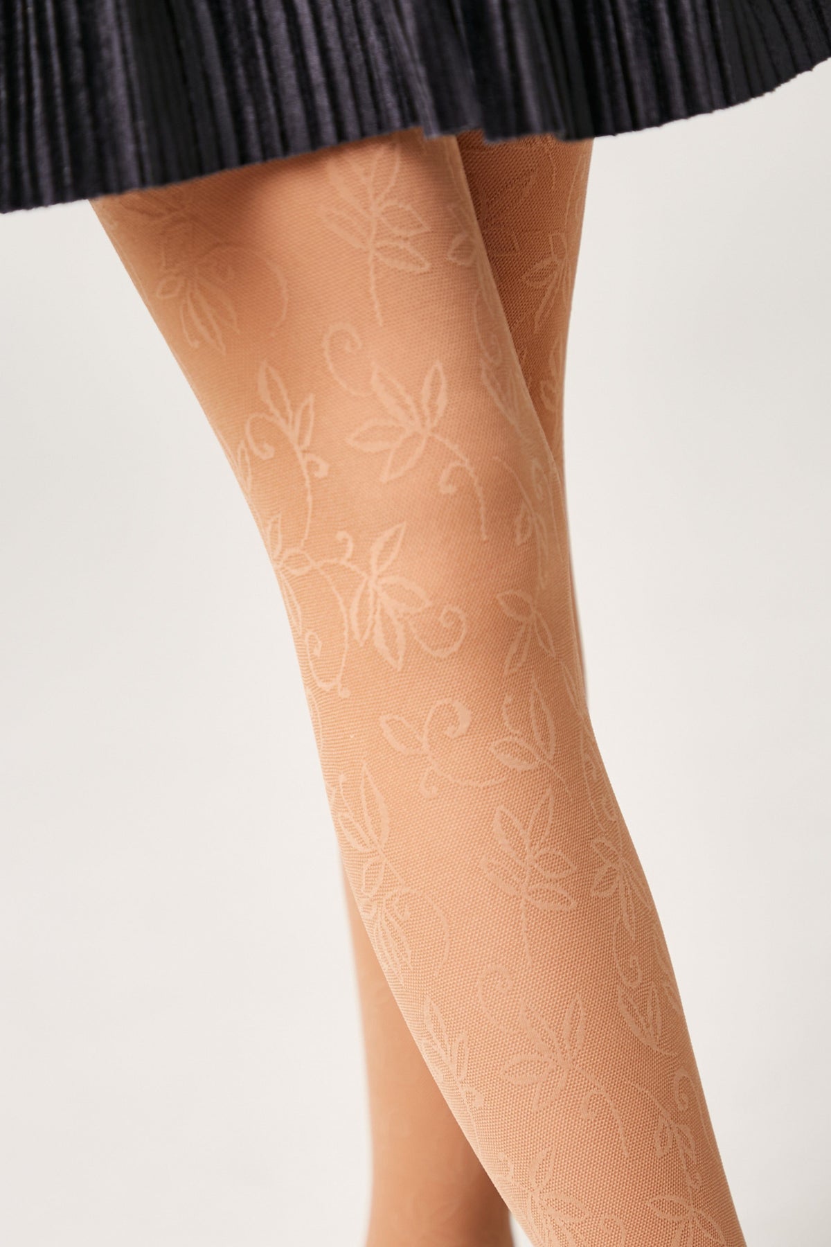 Nude-colored tights featuring a subtle, repeating leaf and vine pattern worn on legs beneath a dark pleated skirt.