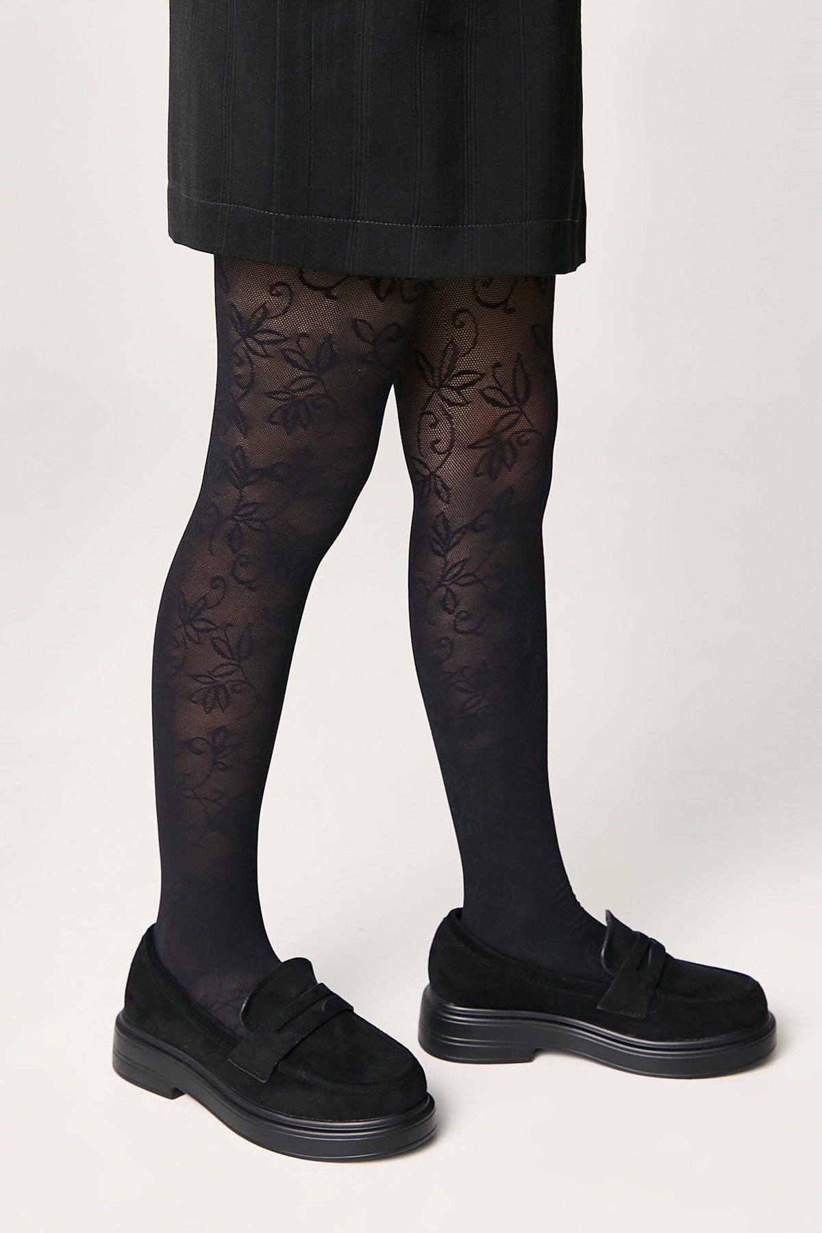 Black patterned tights featuring a delicate floral motif worn with black suede-like loafers and a dark skirt.