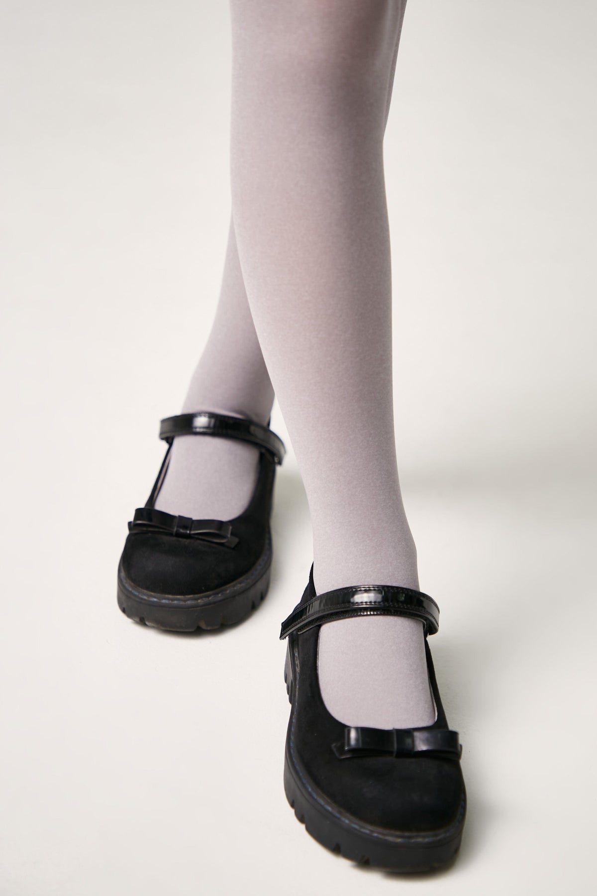 Black mary jane shoes with a bow detail and thick lug soles are worn with light grey opaque tights against a white background.