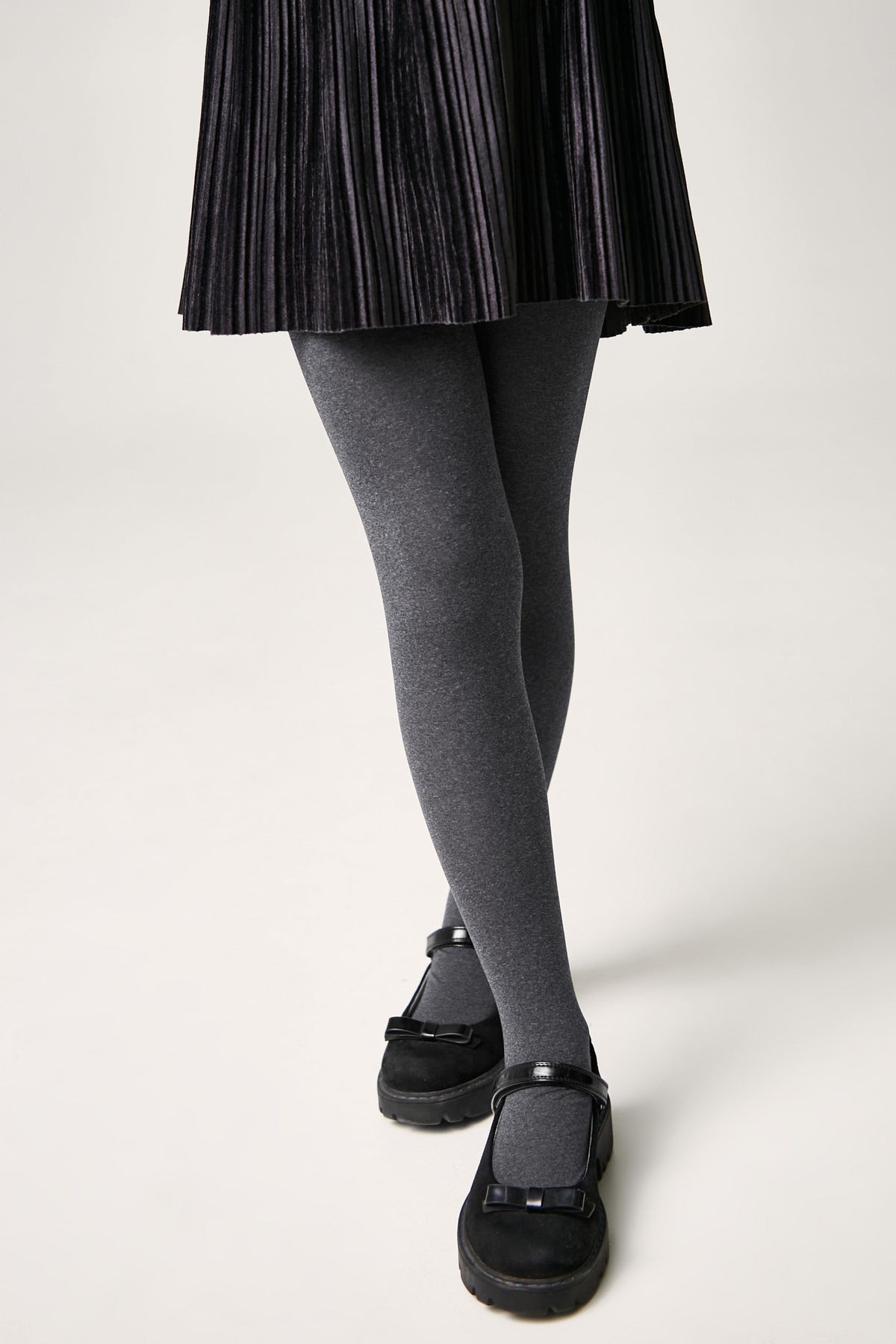 A person wears a black pleated skirt, charcoal grey opaque tights, and black Mary Jane shoes with bow details against a plain background.