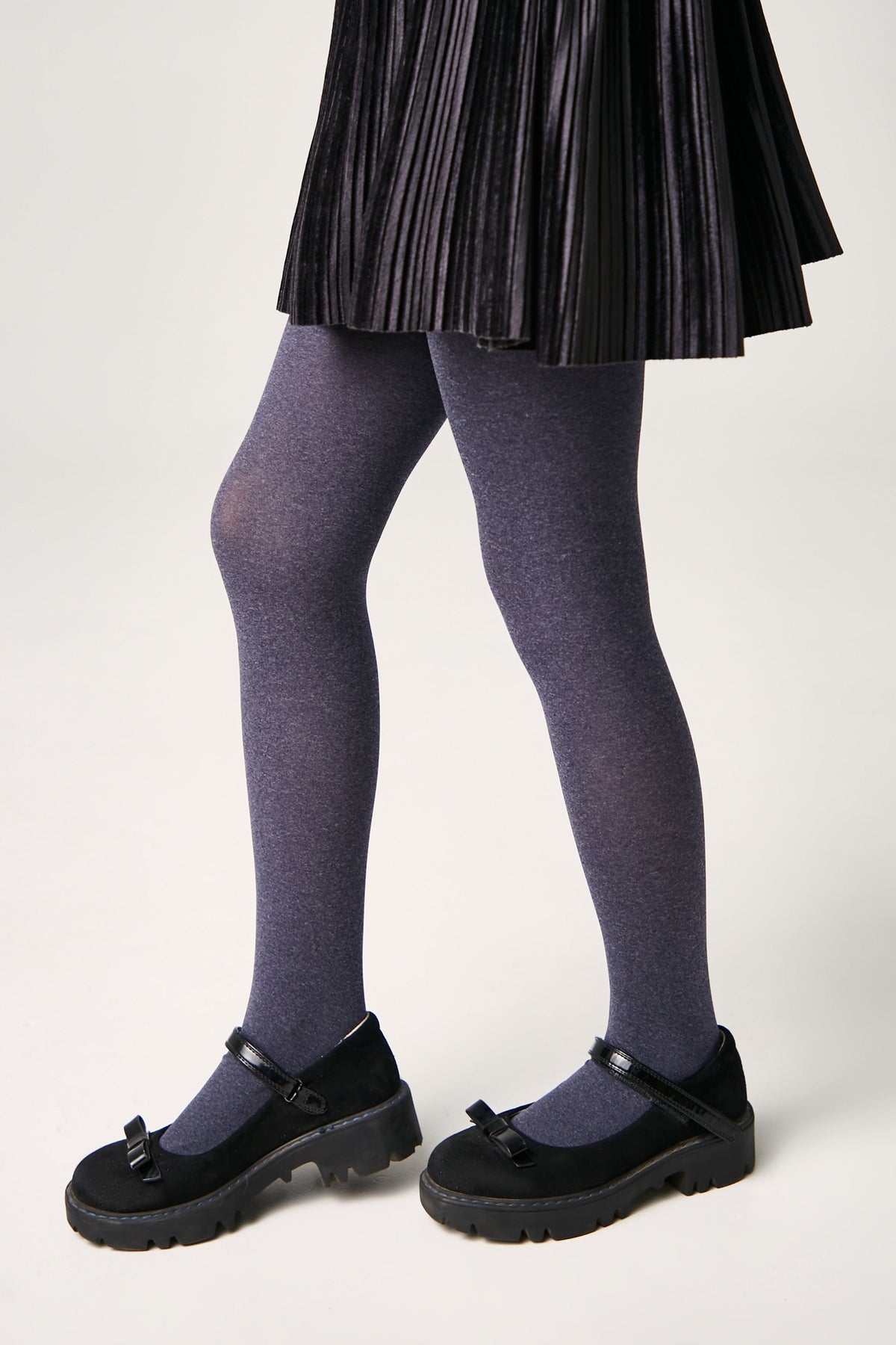 A pair of purple heathered tights worn under a black pleated skirt with black Mary Jane style shoes featuring thick rubber soles and small decorative bows.