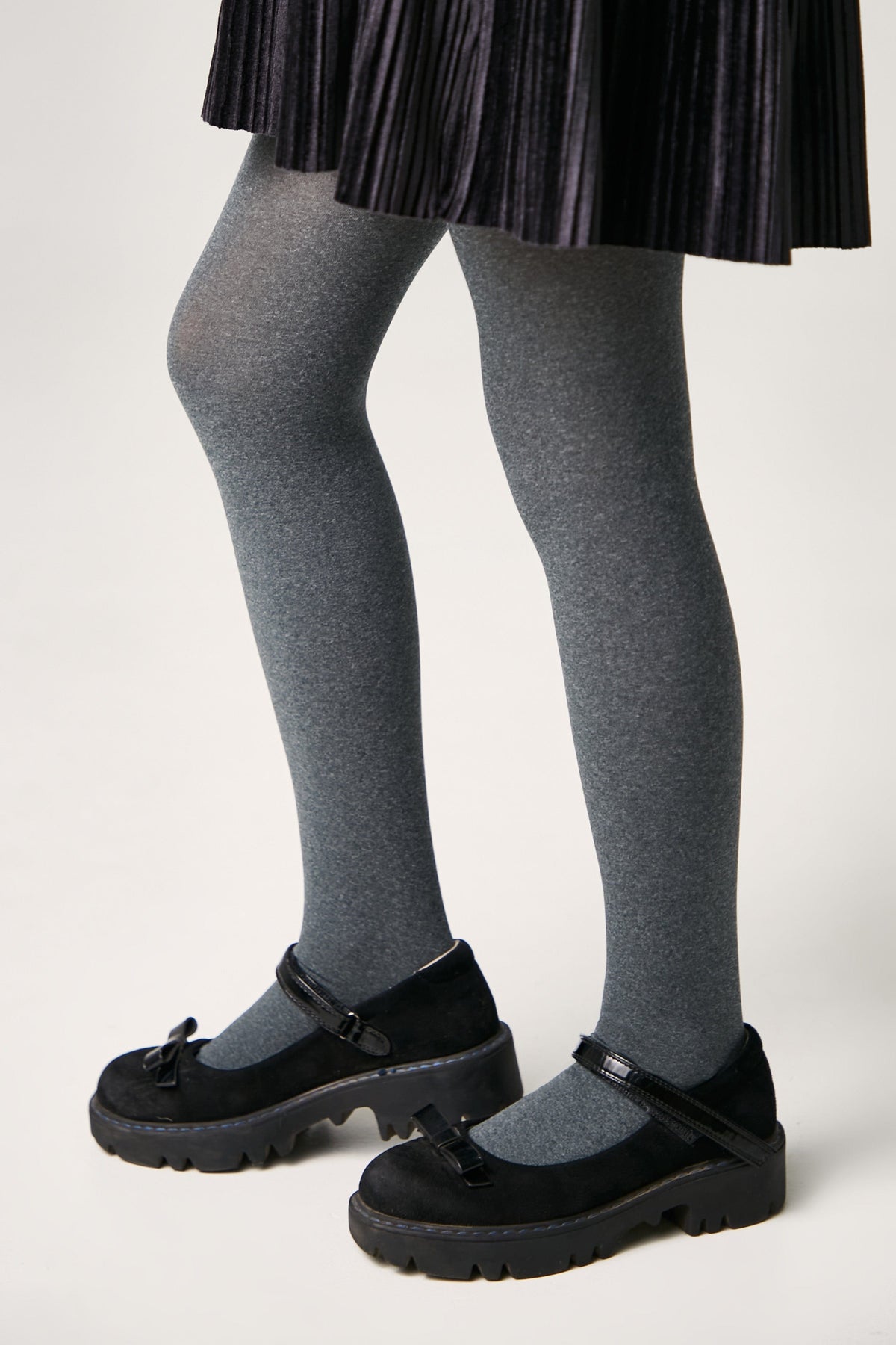 A person wearing a pleated black skirt, grey tights, and black Mary Jane shoes with thick lug soles.