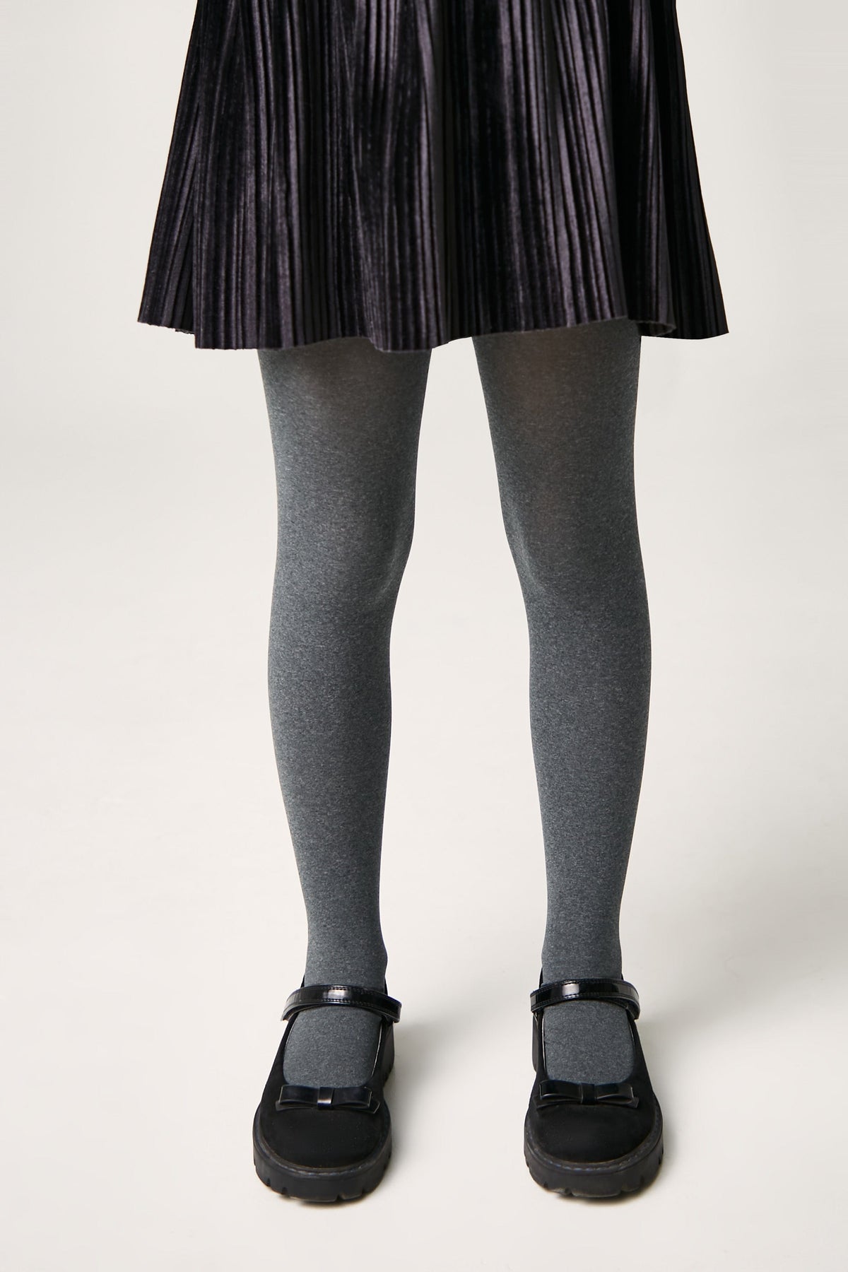 A pair of grey opaque tights worn under a black pleated skirt with black mary jane shoes featuring bows on the front.