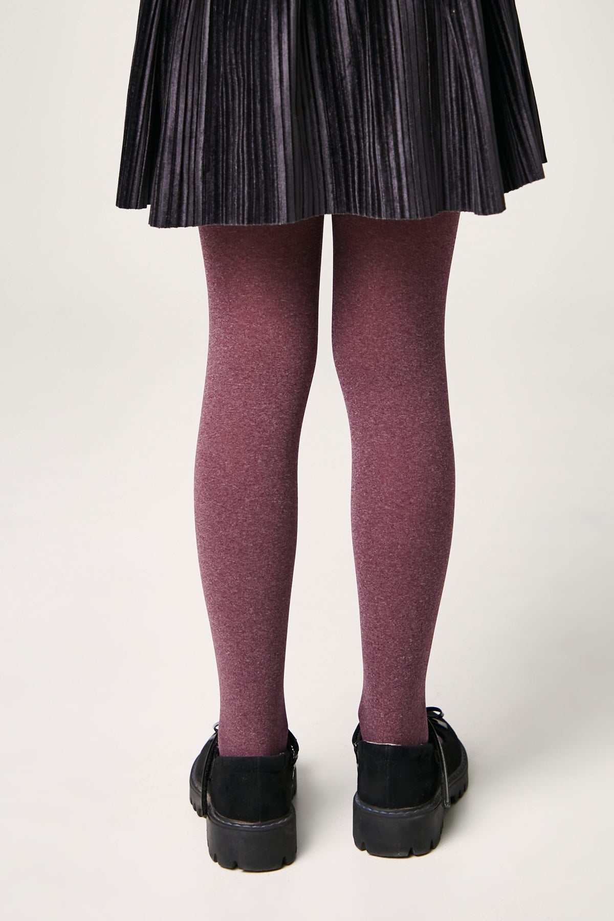A pair of purple heathered tights worn by a child with a black pleated skirt and black flat shoes.