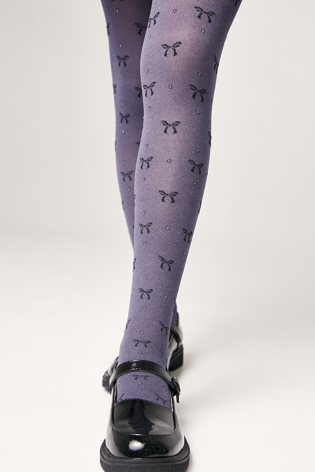 Grey tights featuring a repeating pattern of small dark bows and subtle glitter accents are worn with black patent leather Mary Jane shoes.