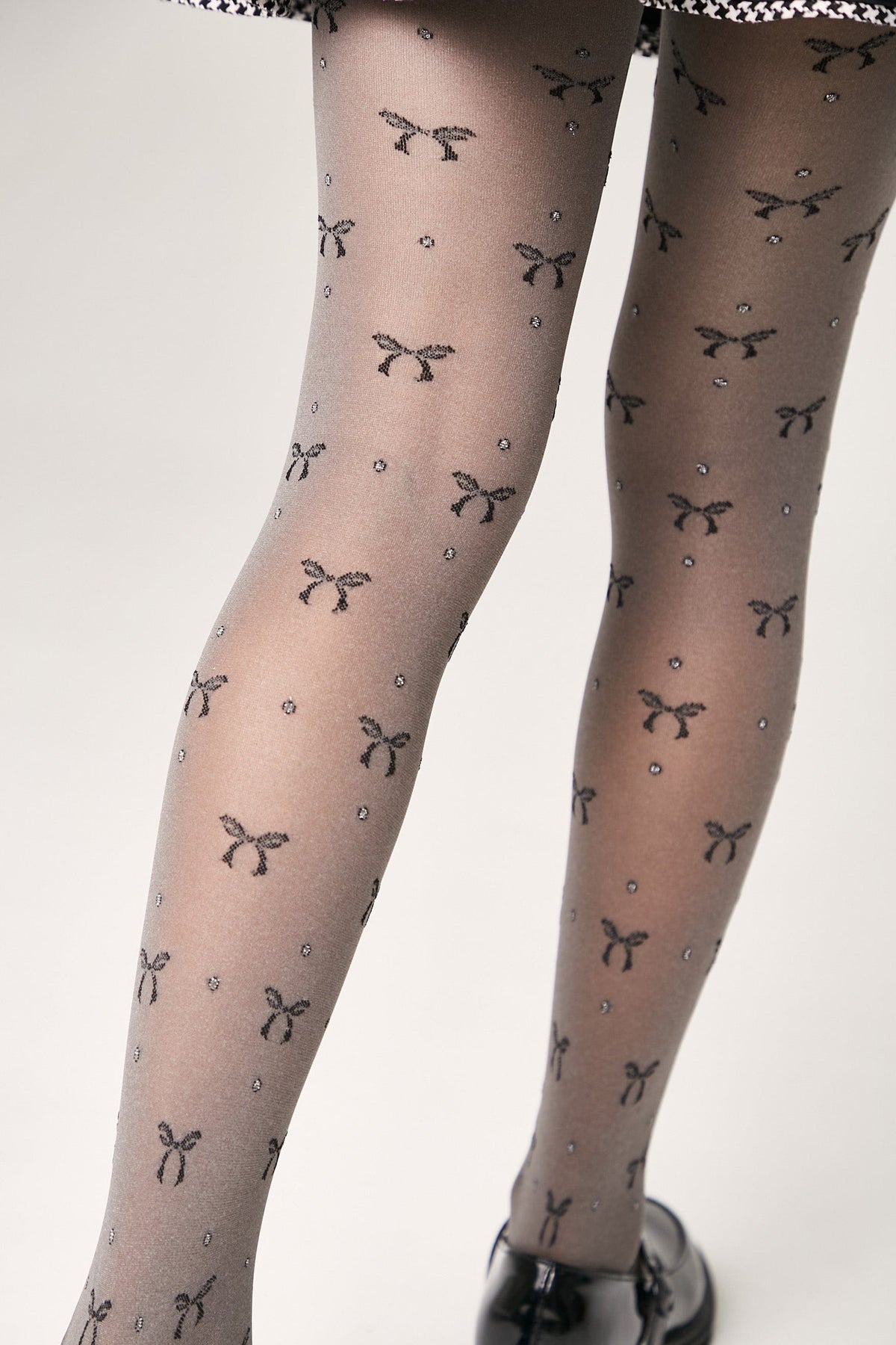 Sheer grey tights patterned with a repeating print of small black bows and tiny sparkling dots, worn with black shoes.