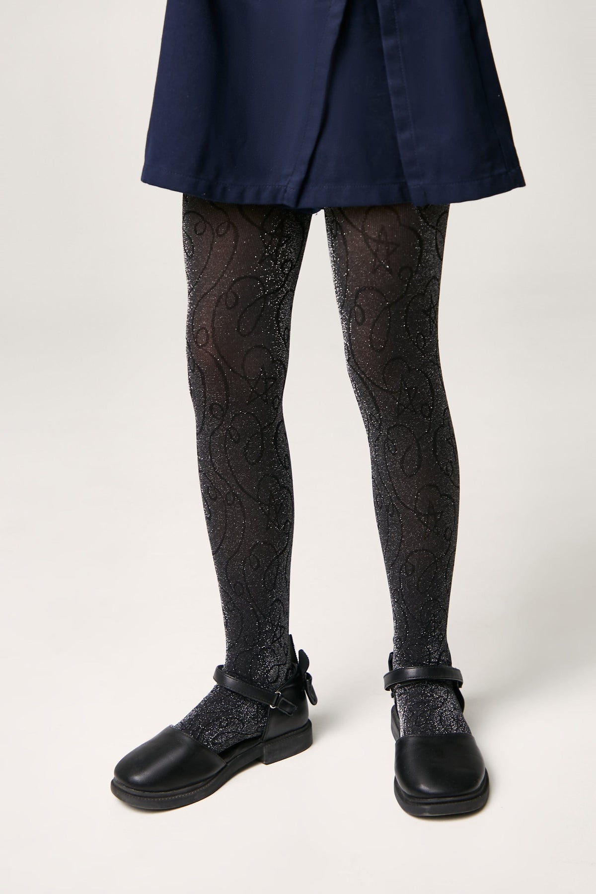 A pair of black glittery tights featuring a subtle swirling pattern worn under a navy blue skirt with black buckled flat shoes.