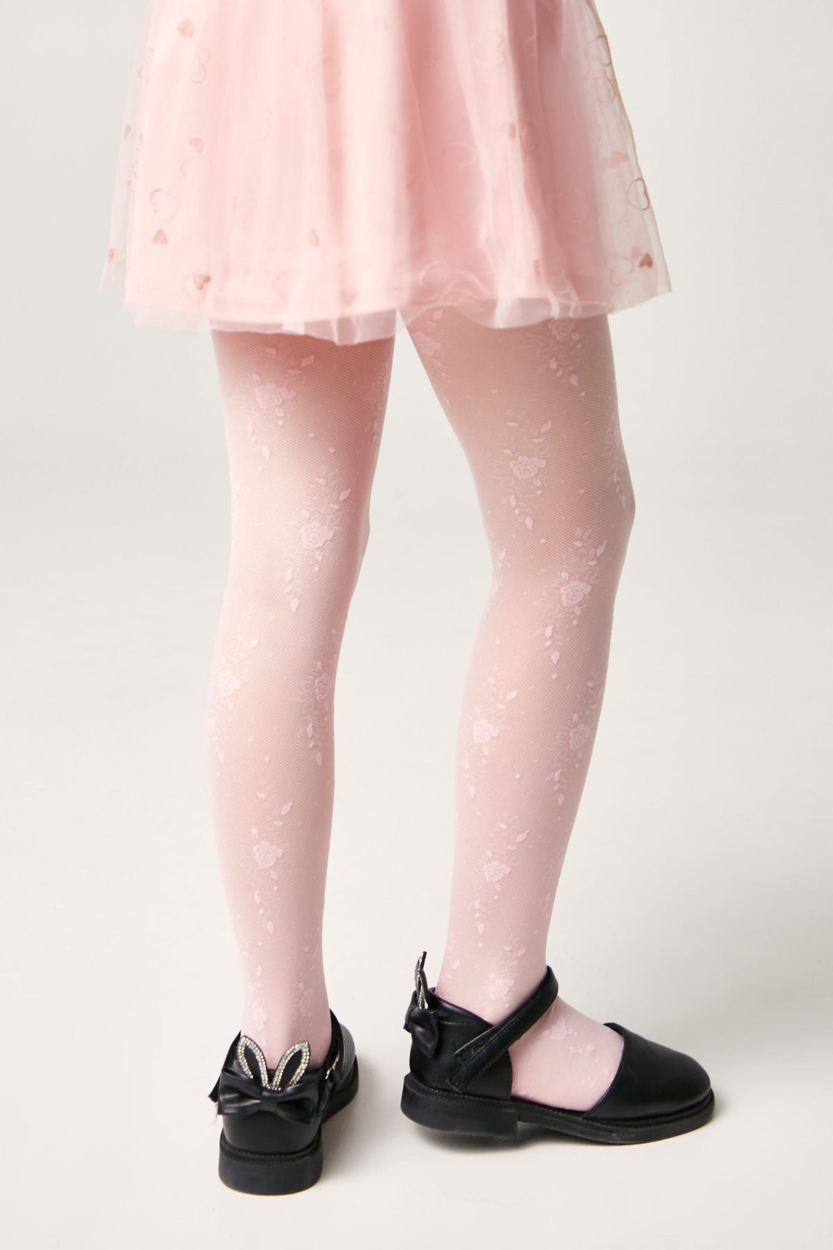 A pair of light pink floral patterned tights worn with black Mary Jane shoes featuring small rabbit ears and bows on the heels.