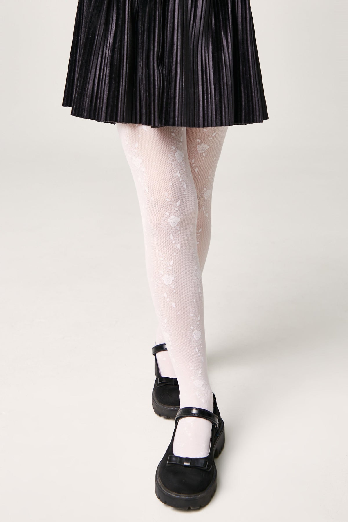 A pair of sheer white floral patterned tights worn with a black pleated skirt and black Mary Jane shoes against a white background.