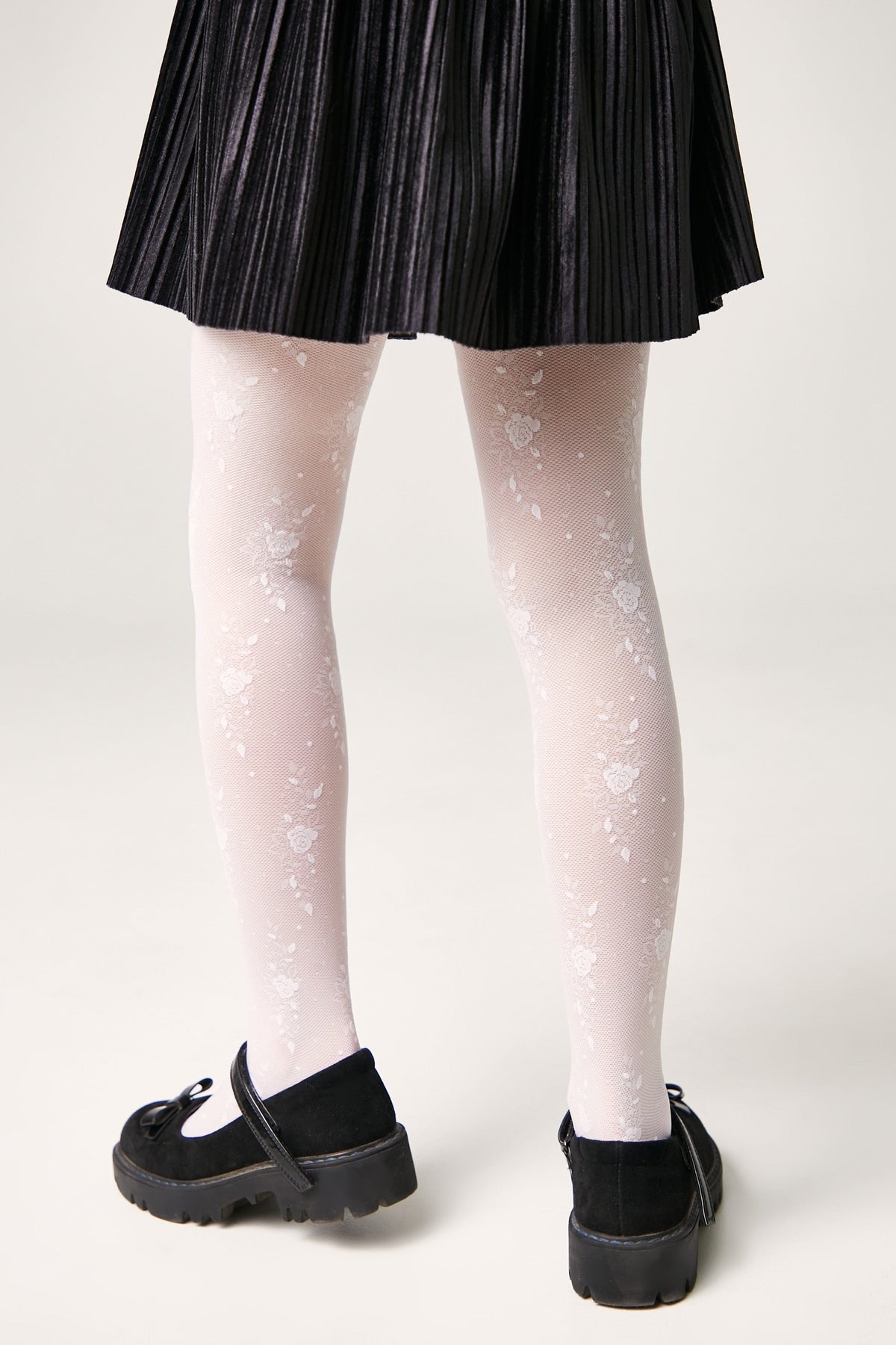 A person wears white floral-patterned tights underneath a black pleated skirt paired with black Mary Jane shoes.