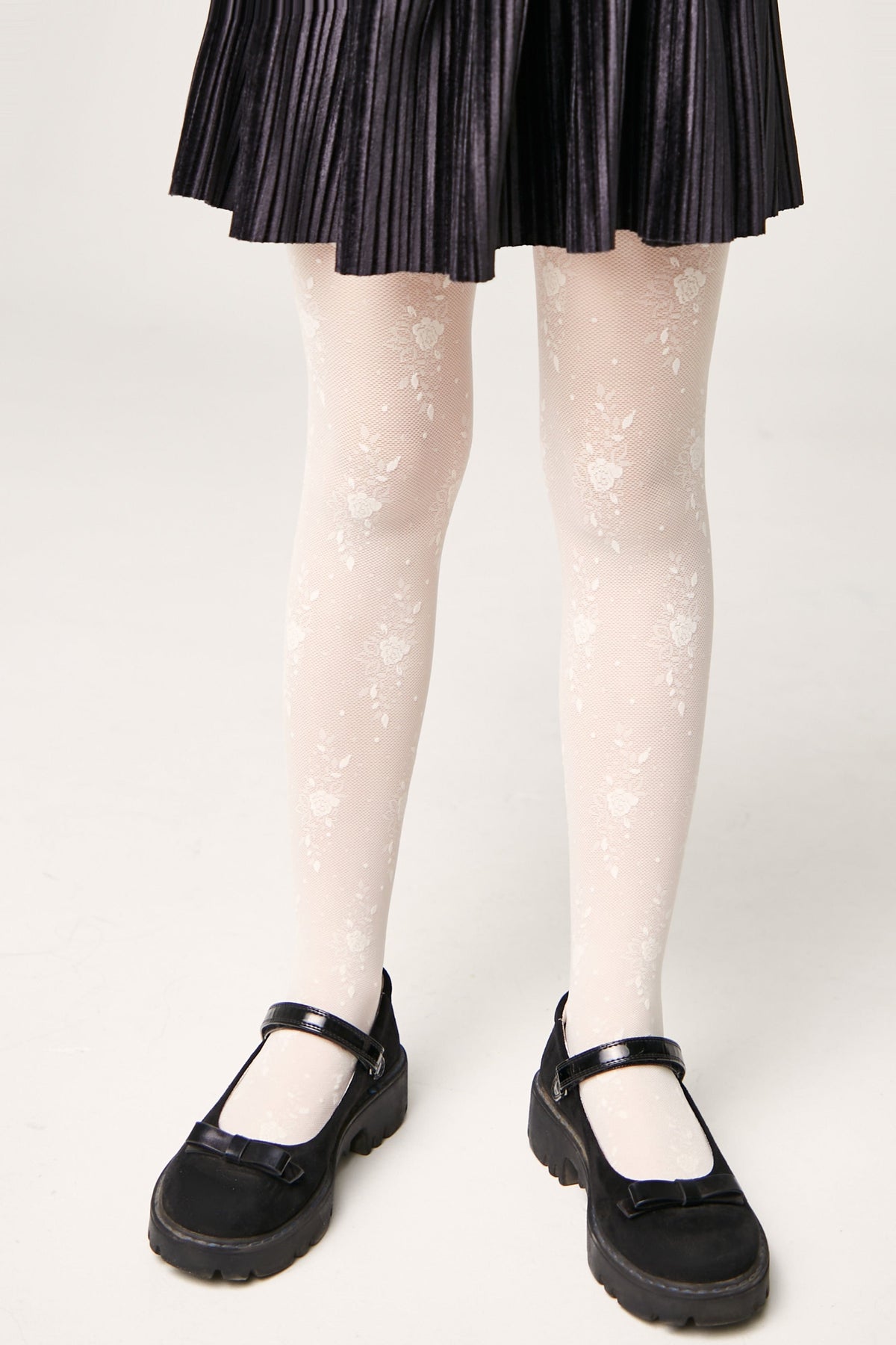 White patterned tights with a delicate floral design worn with a black pleated skirt and black Mary Jane shoes against a white background.