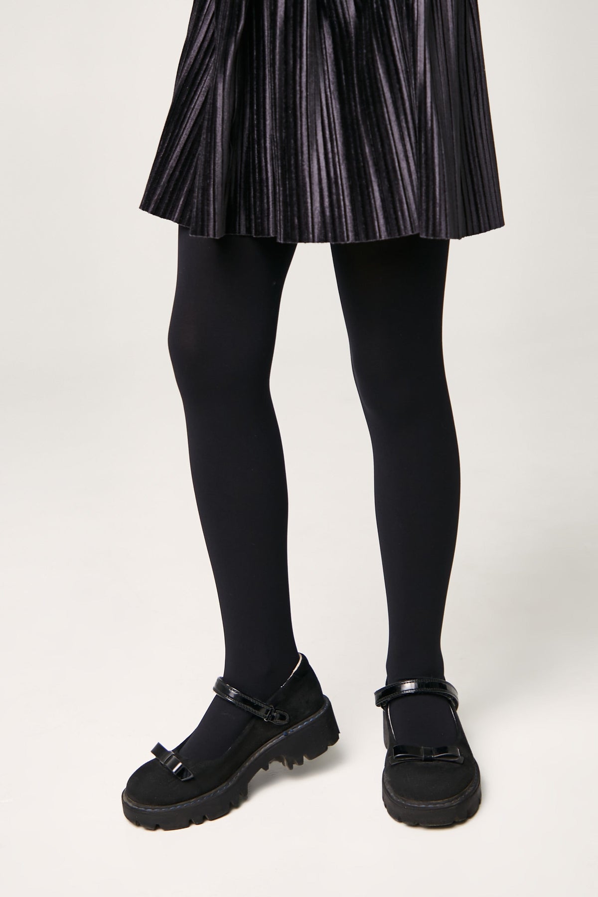 Black Mary Jane style shoes featuring a bow detail on the toe, chunky lug soles, and adjustable ankle straps worn with a pleated black skirt and opaque tights.