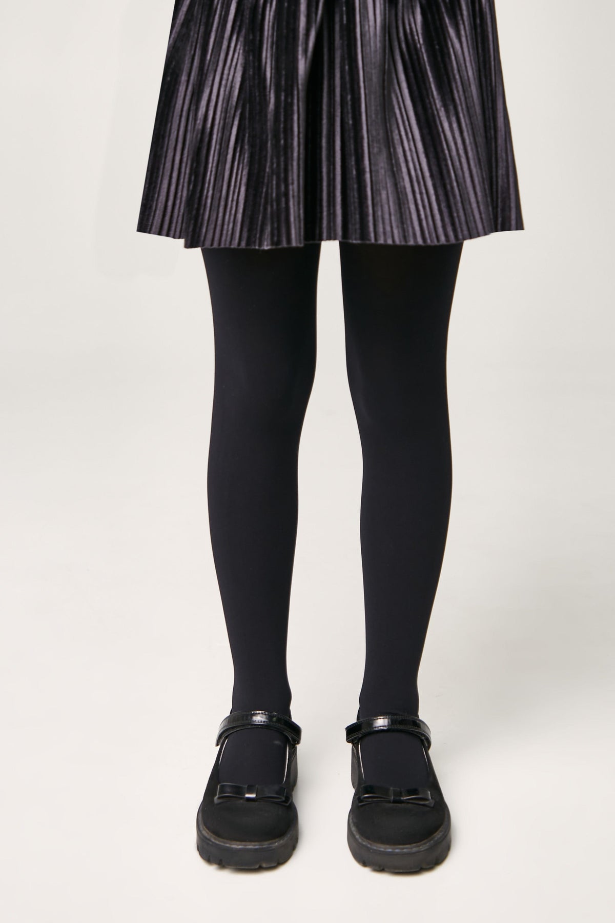 A person wears a dark grey pleated skirt with solid black opaque tights and black flat shoes featuring a bow detail on the toe.